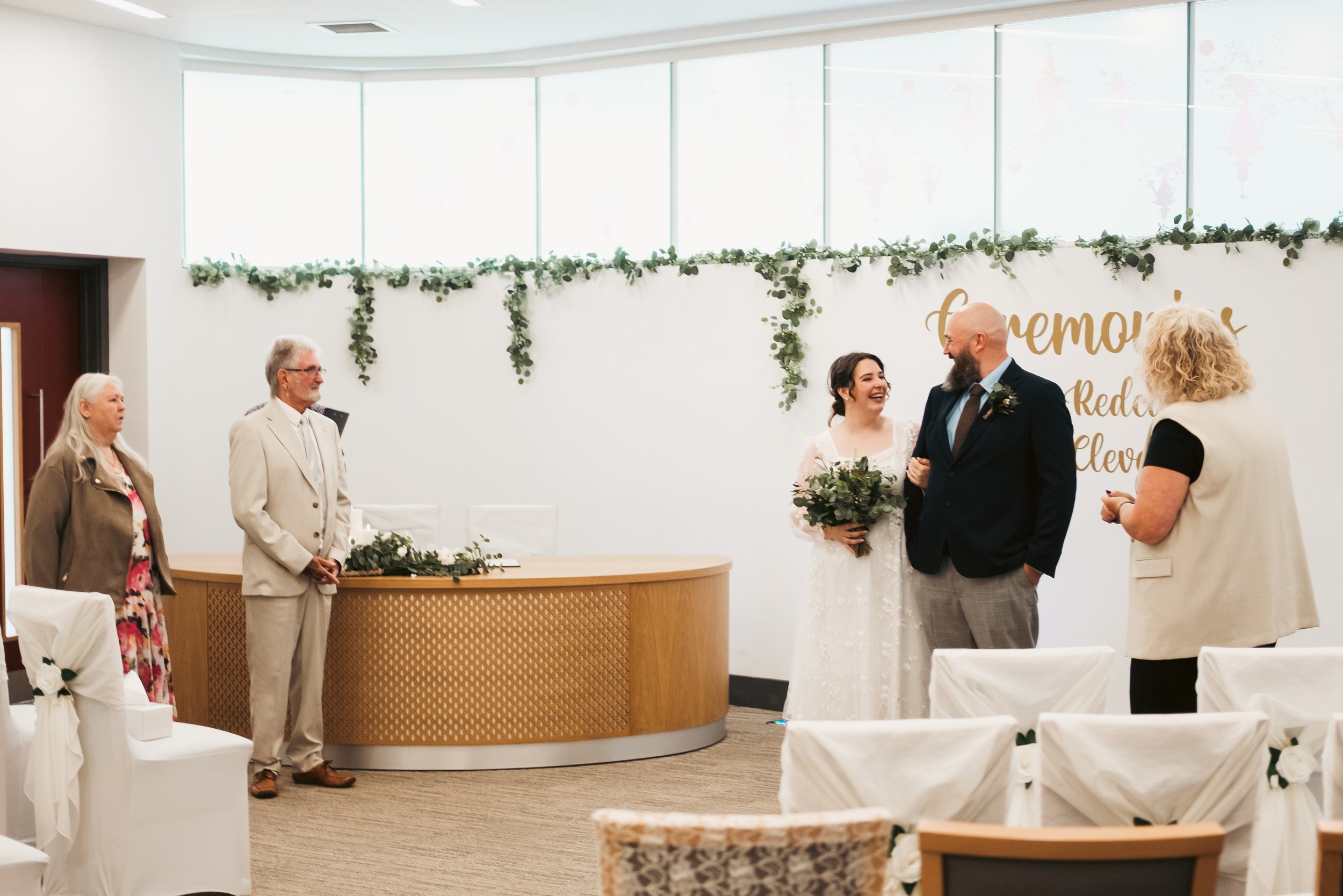 A bride and groom smiling and holding hands during a wedding ceremony, with guests and floral decorations in the background.
