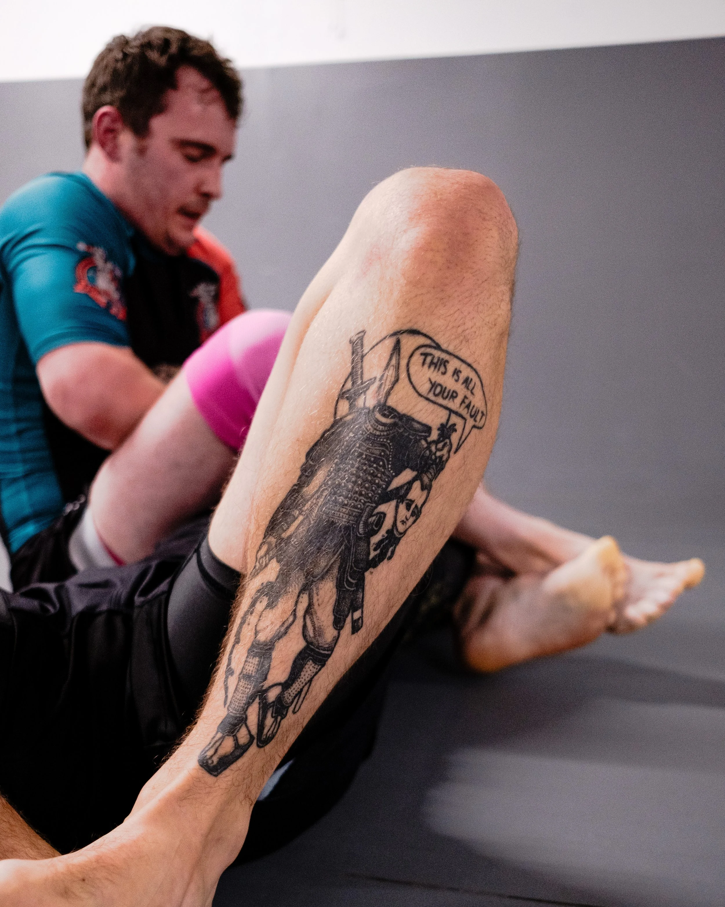 Close-up of a tattoo on a person's leg featuring a warrior figure with a speech bubble that says, 'This is all your fault.'