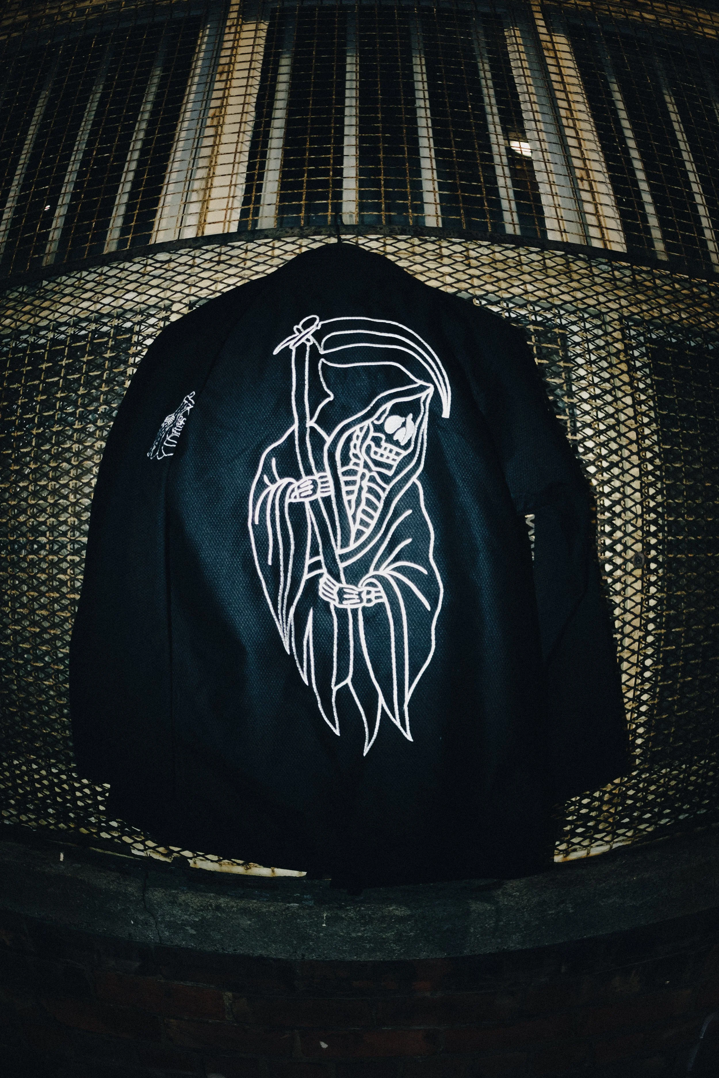 A black jacket with a white embroidered skeleton figure dressed as a Grim Reaper, holding a scythe, hanging on a metal mesh rack.