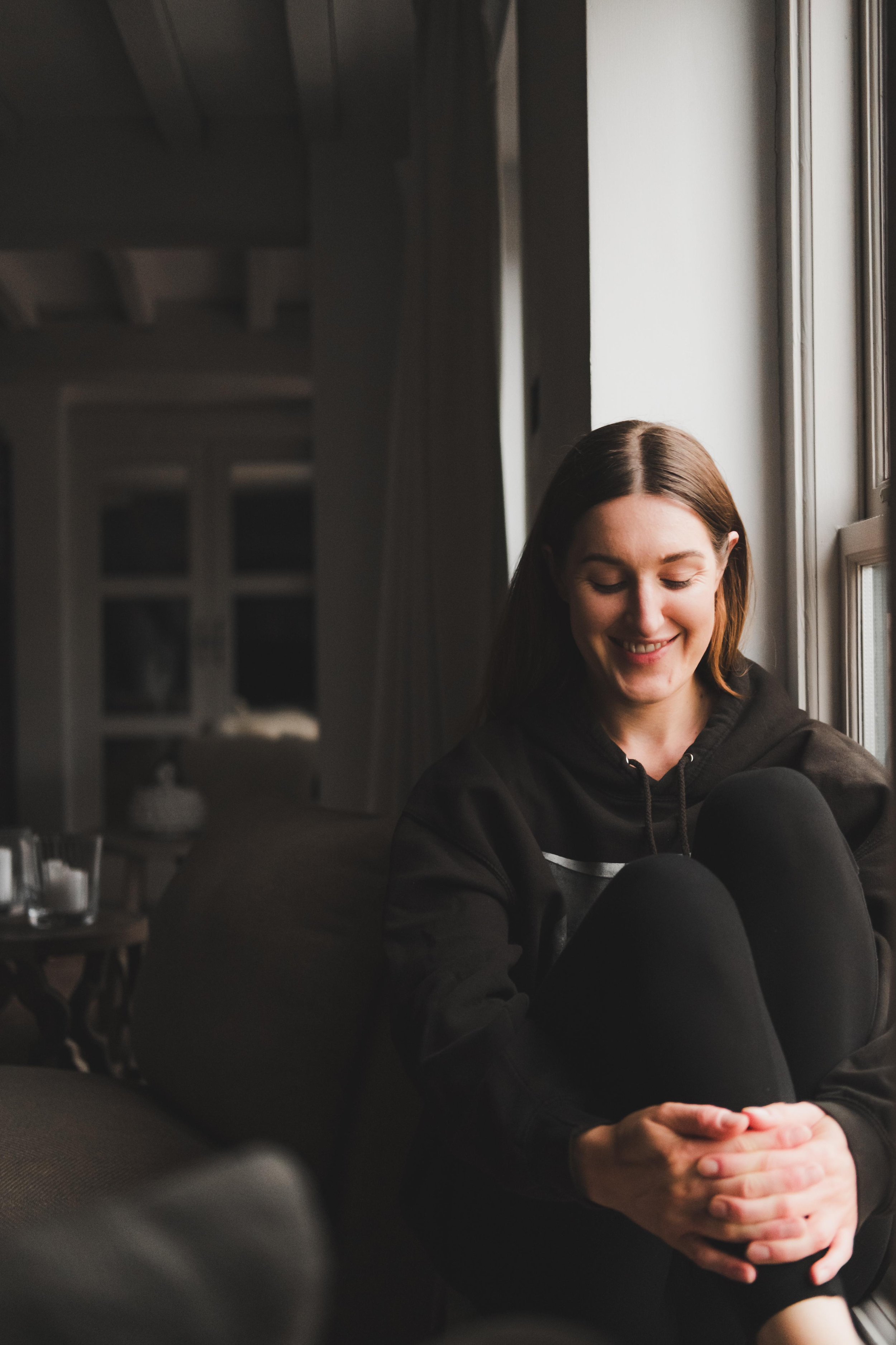 A young woman with brown hair sitting by a window in a cozy room, smiling with her eyes closed, wearing a black hoodie, with her hands clasped together in her lap.