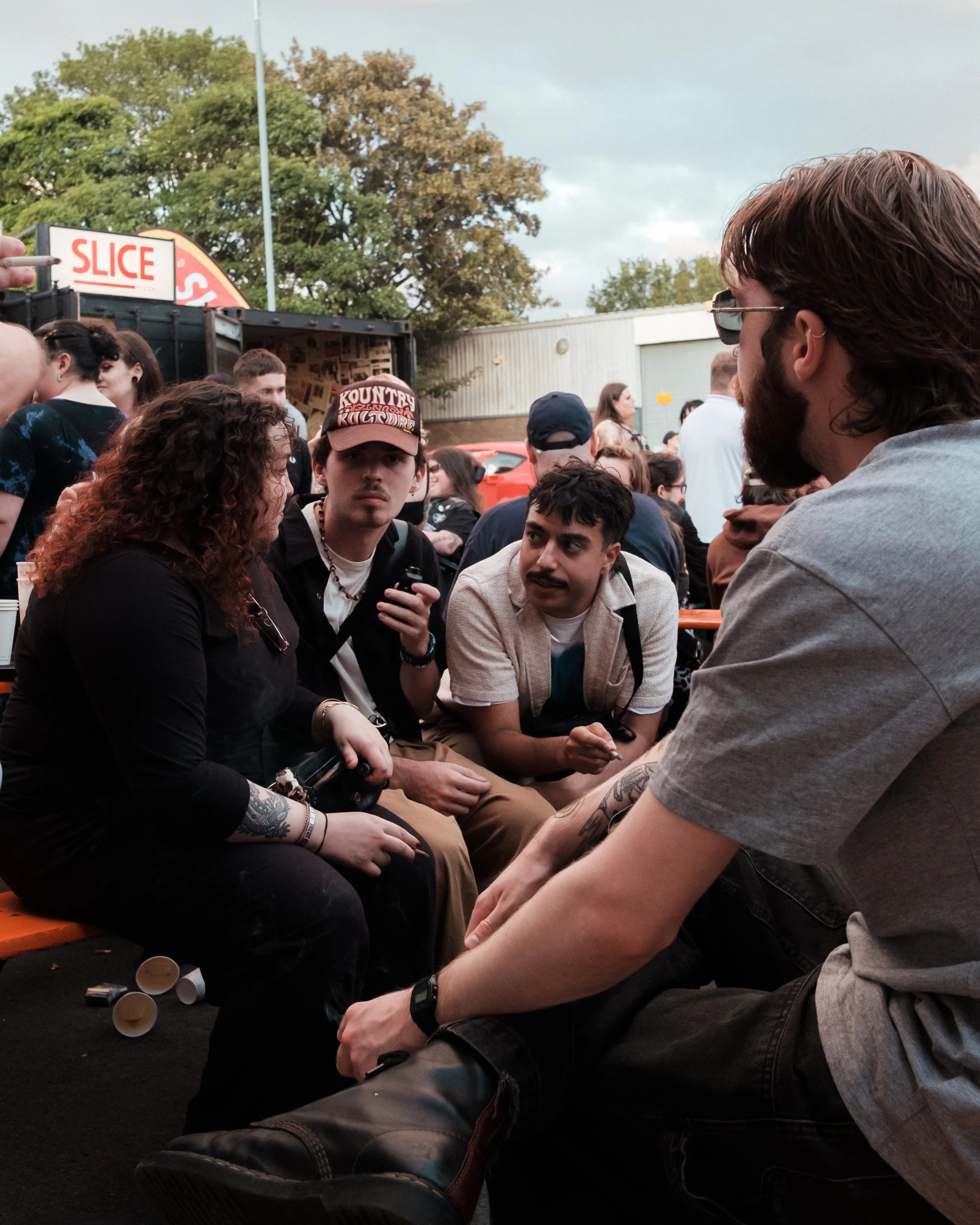 Group of young adults sitting outdoors at a social gathering or festival, engaged in conversation. There are food trucks and trees in the background.