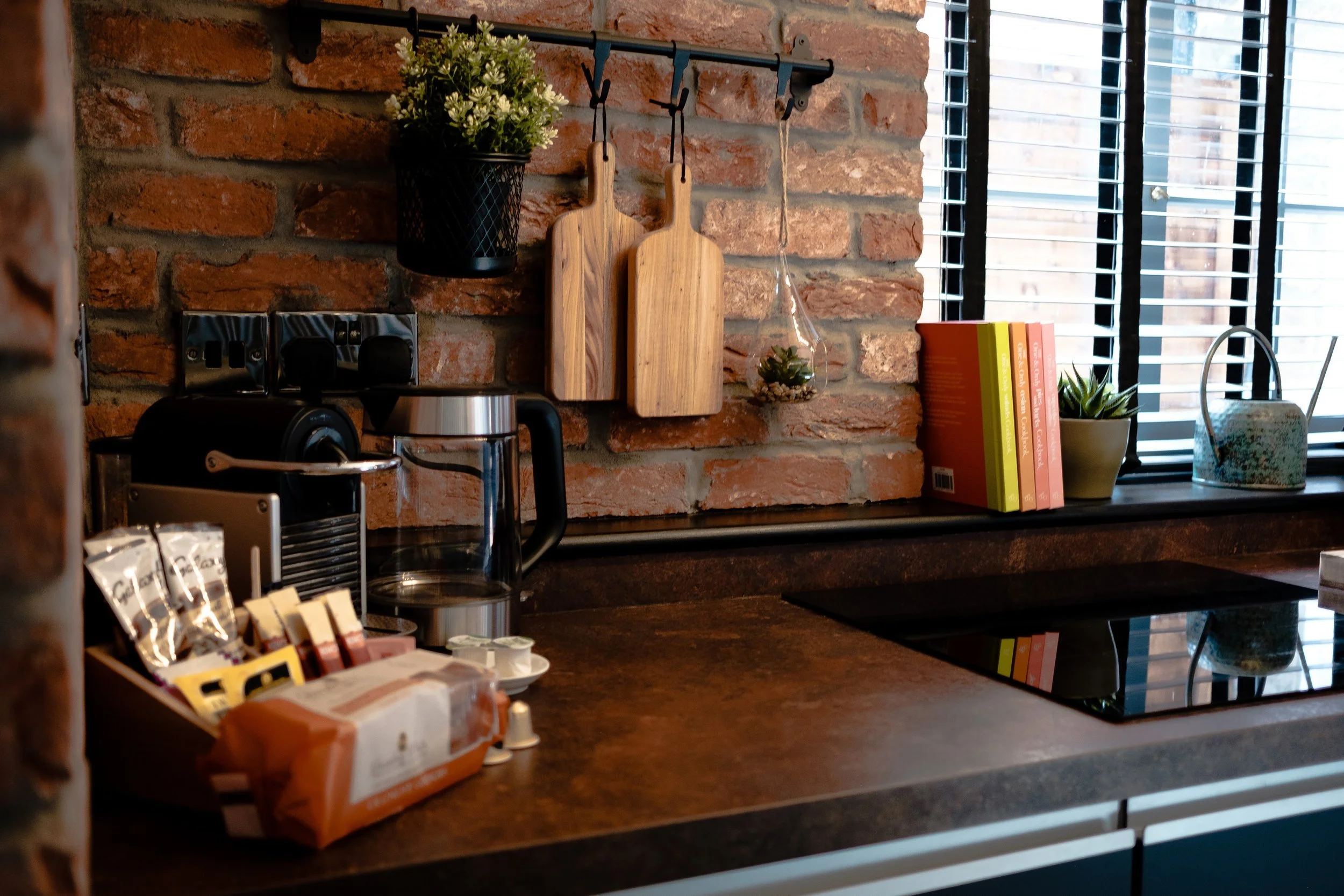 A kitchen countertop with a coffee machine, jars, and snacks in the foreground, a brick wall with two cutting boards, a plant, and books on the windowsill in the background.