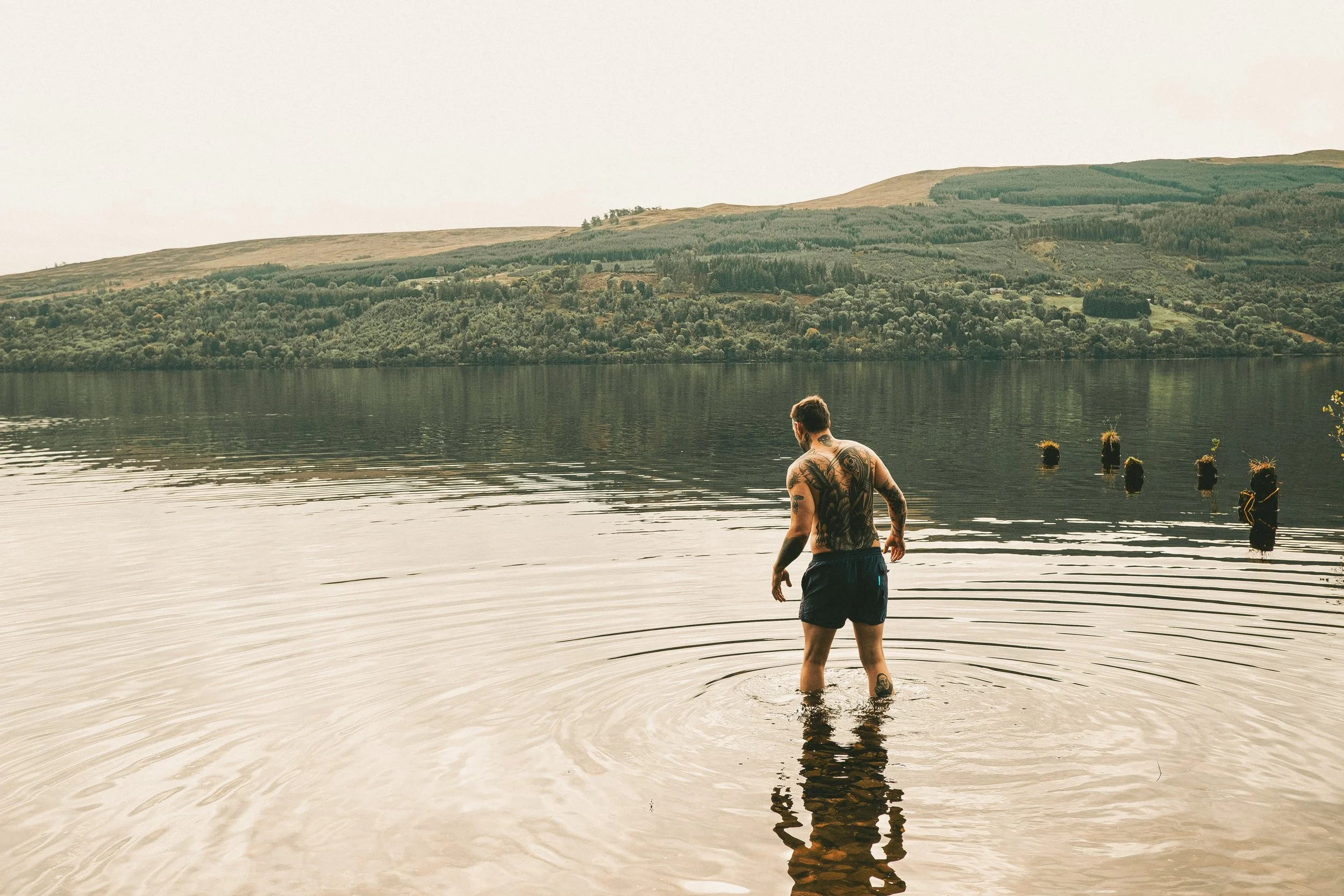 A man with tattoos in black shorts standing in a calm lake, facing away and looking towards hills in the background.