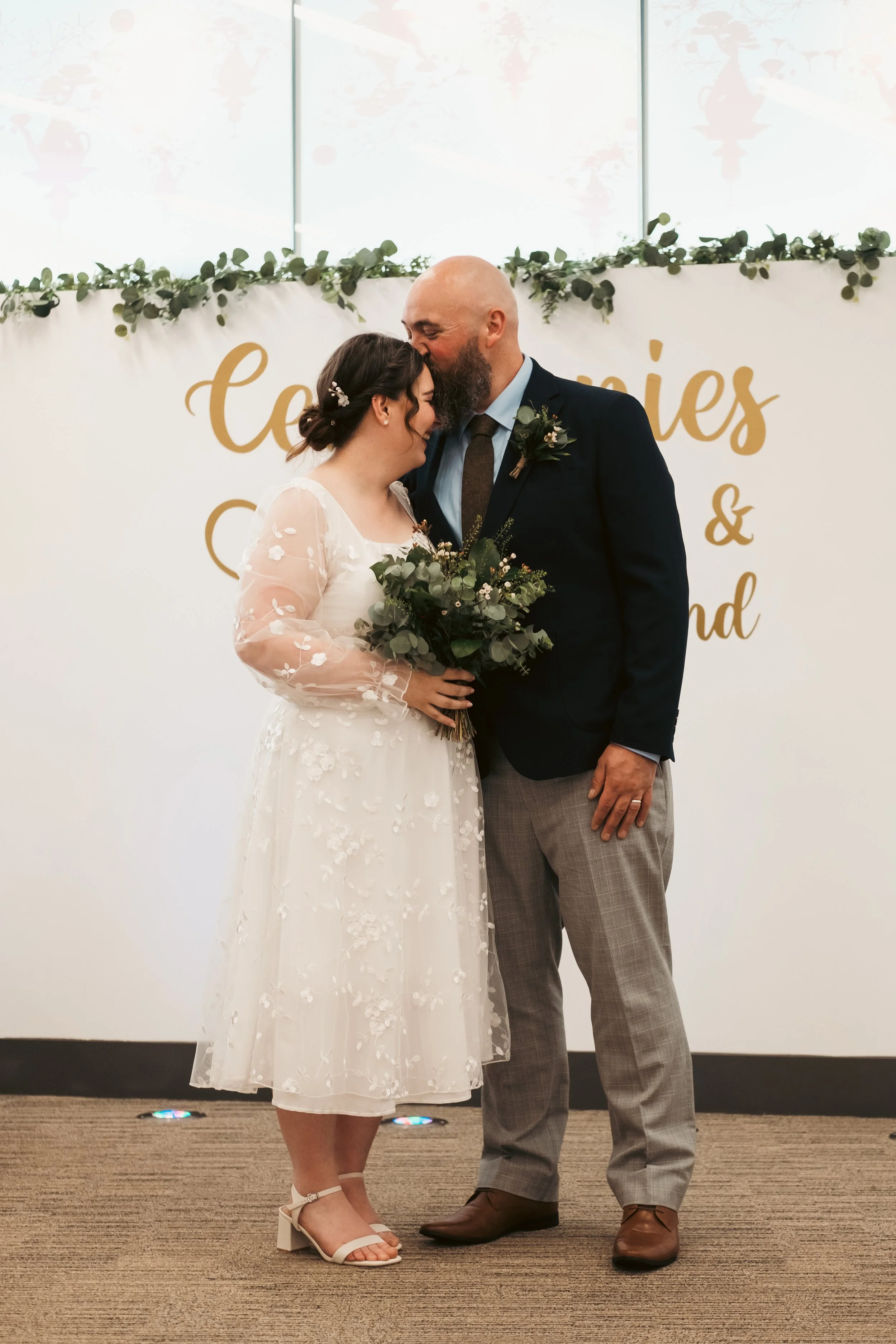 A bride and groom sharing a moment during their wedding ceremony. The bride is wearing a white dress with floral embroidery and the groom is in a dark jacket and plaid trousers. They are standing in front of a decorated backdrop with foliage and a si