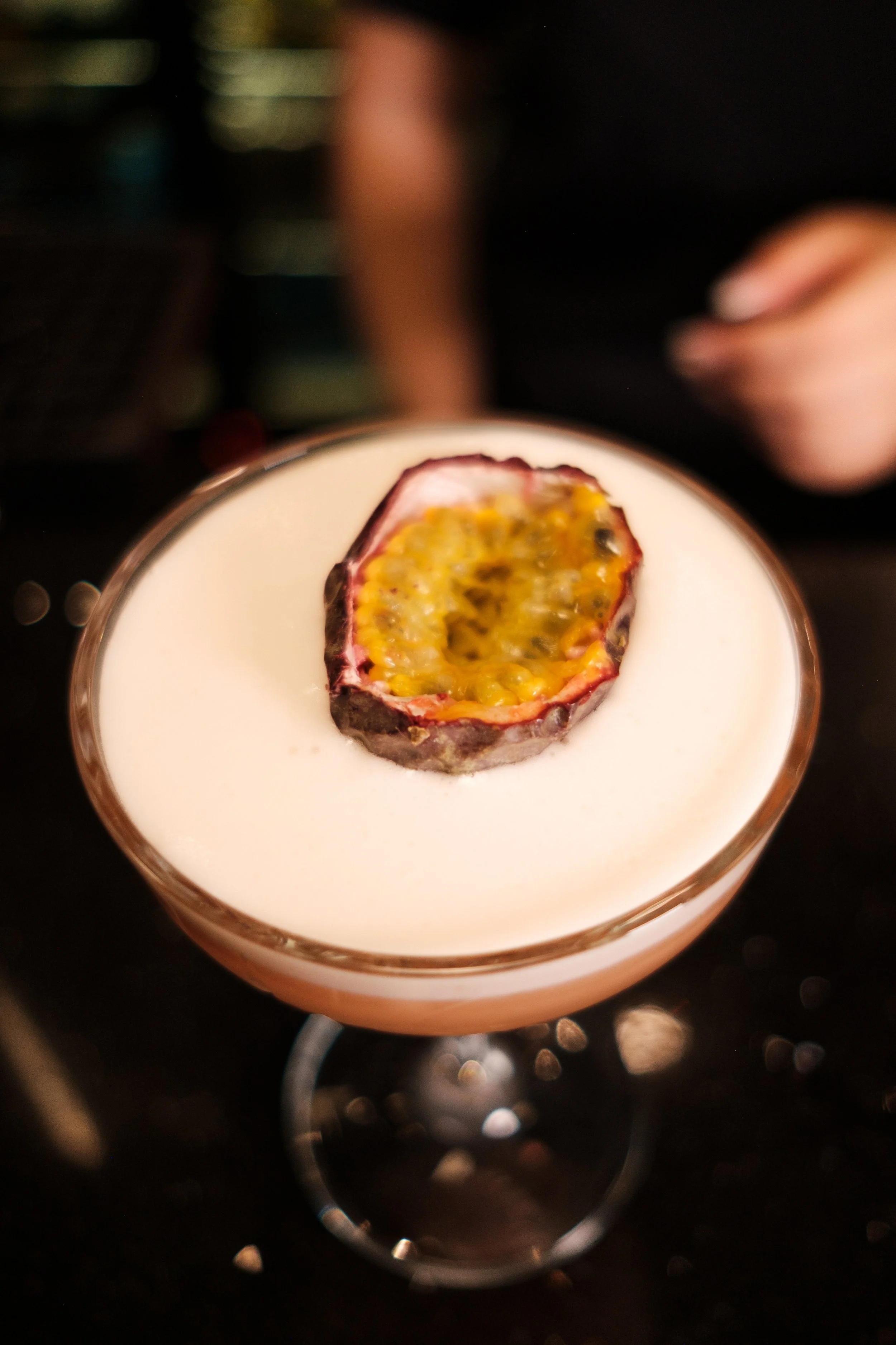A close-up of a passion fruit half on top of a creamy white drink. The passion fruit has yellow and black seeds inside. The background is dark and blurry.