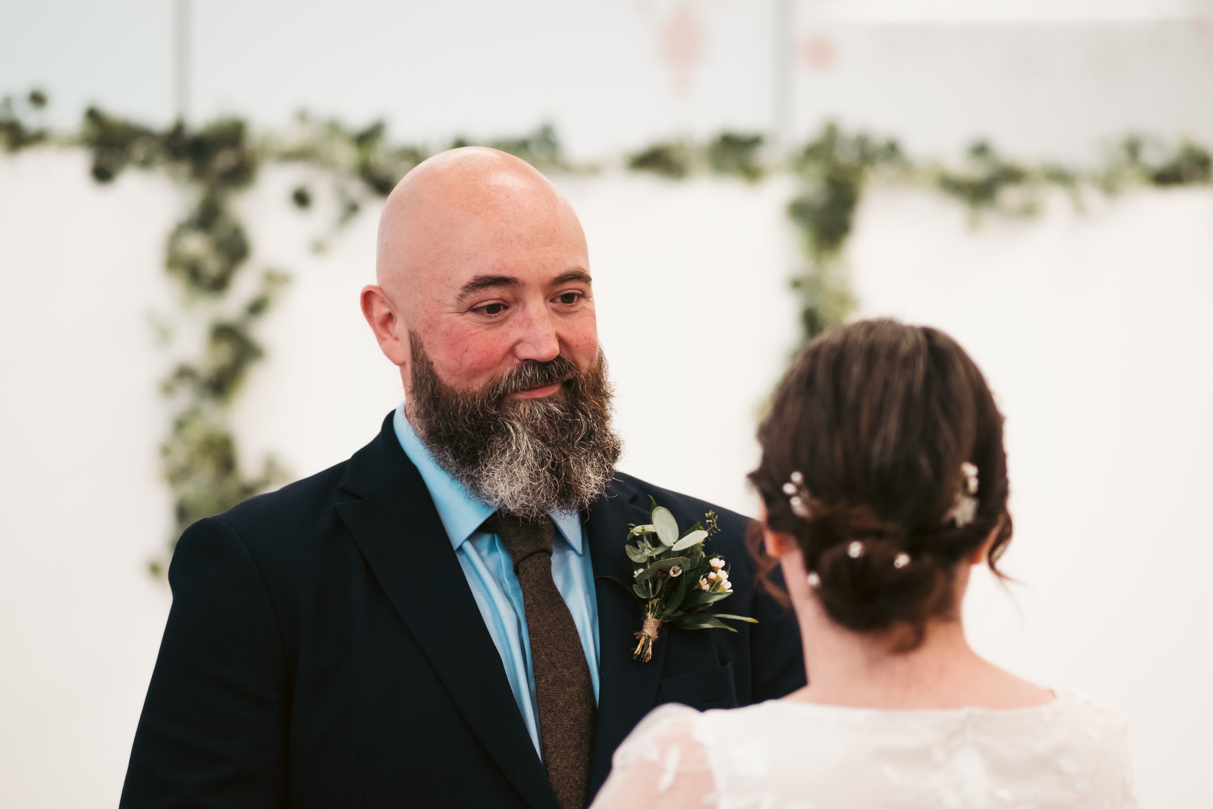 A man with a beard and bald head wearing a dark suit and blue shirt, standing in front of a woman with dark hair in a bun, during a wedding ceremony.