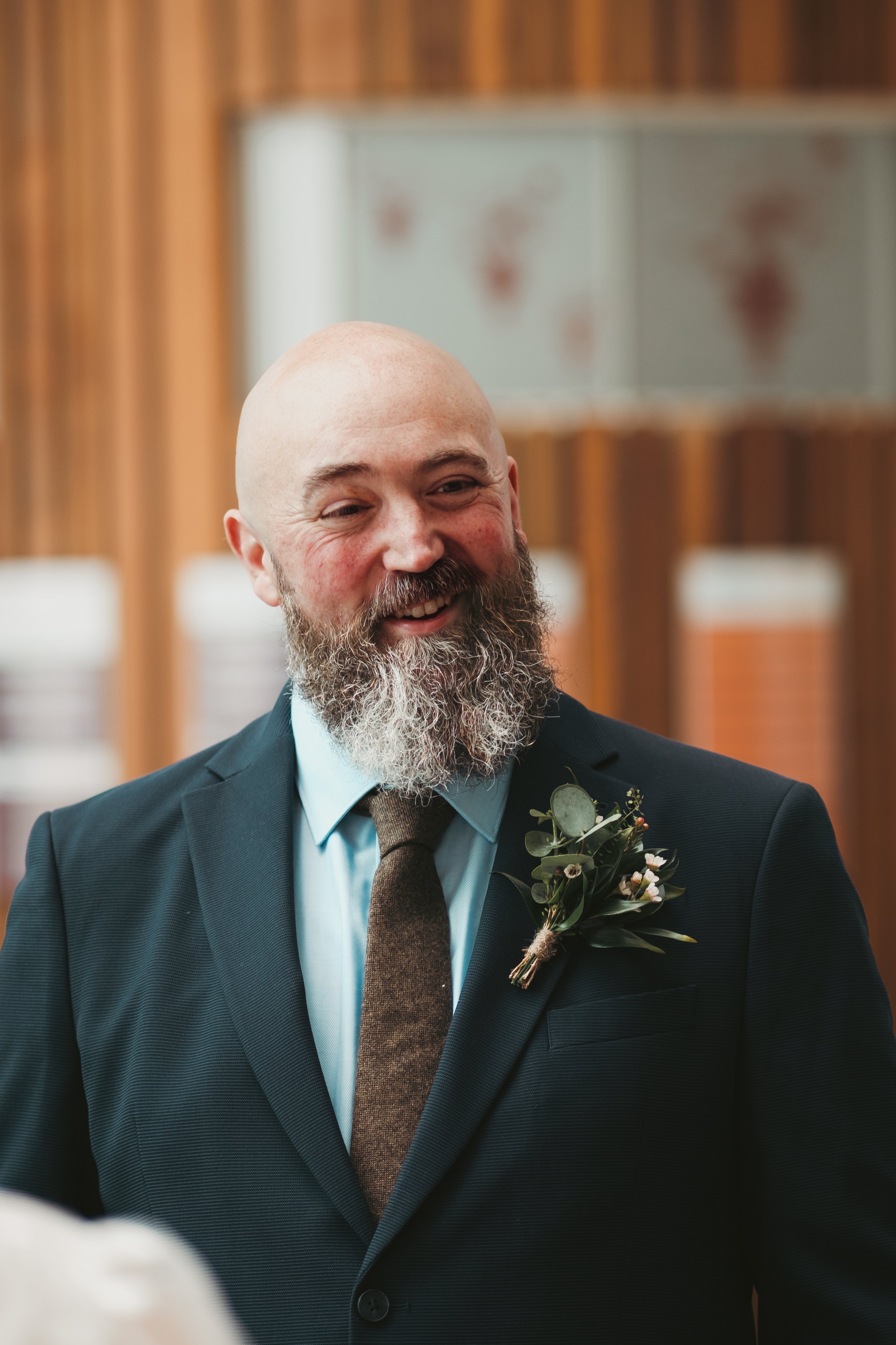 A smiling man with a beard and a bald head wearing a dark suit, light blue shirt, and brown tie, with a boutonnière made of greenery and small flowers, in a room with wooden walls.