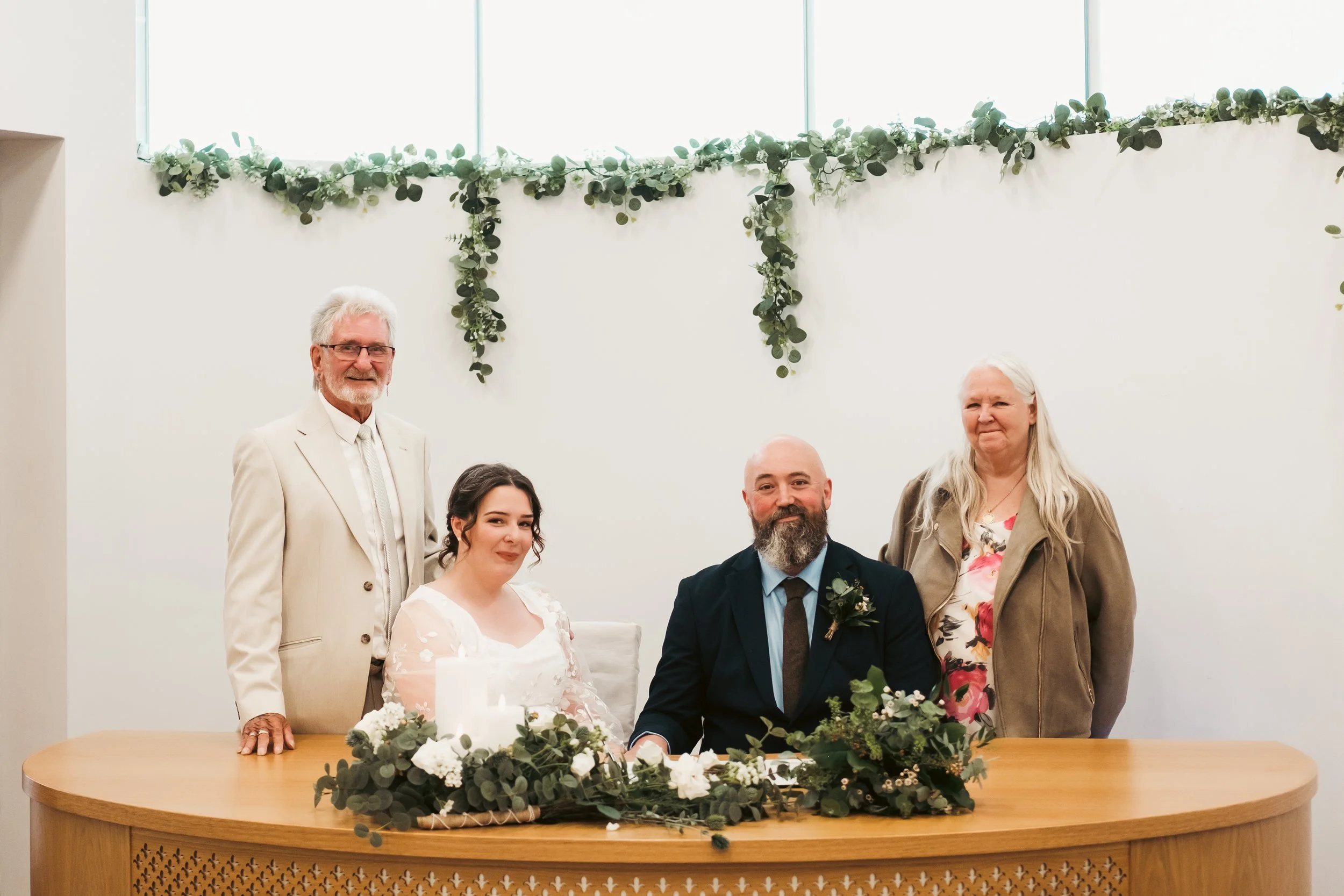 Group of six people celebrating at a wedding reception, including bride and groom seated at a table decorated with white flowers and greenery, and four other guests standing behind them, in a bright room with a white wall and a green draped garland d