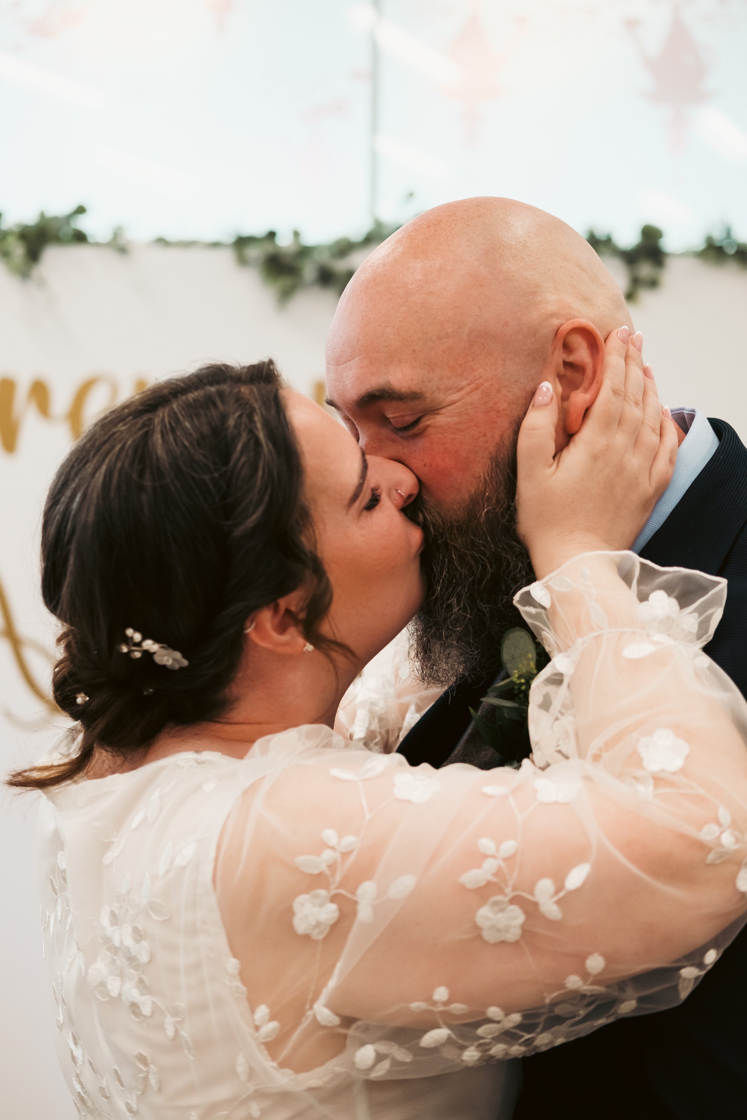 A couple sharing a kiss at their wedding, with the bride in a sheer lace dress and the groom in a suit.