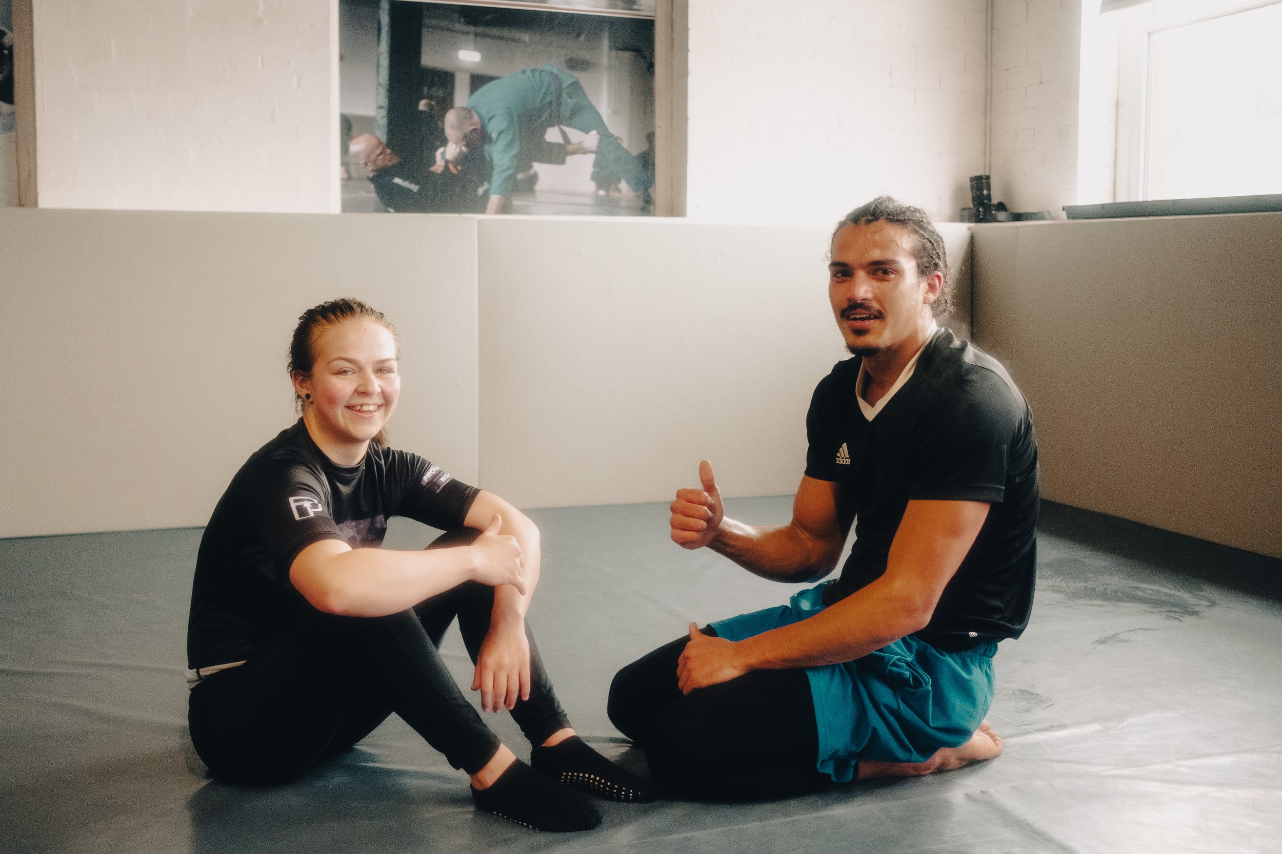 A woman and a man practicing Brazilian Jiu-Jitsu. The woman is sitting on the ground with her arm flexed, smiling at the camera. The man is kneeling beside her, giving a thumbs-up. They are in a gym with padded mats. In the background, two people are