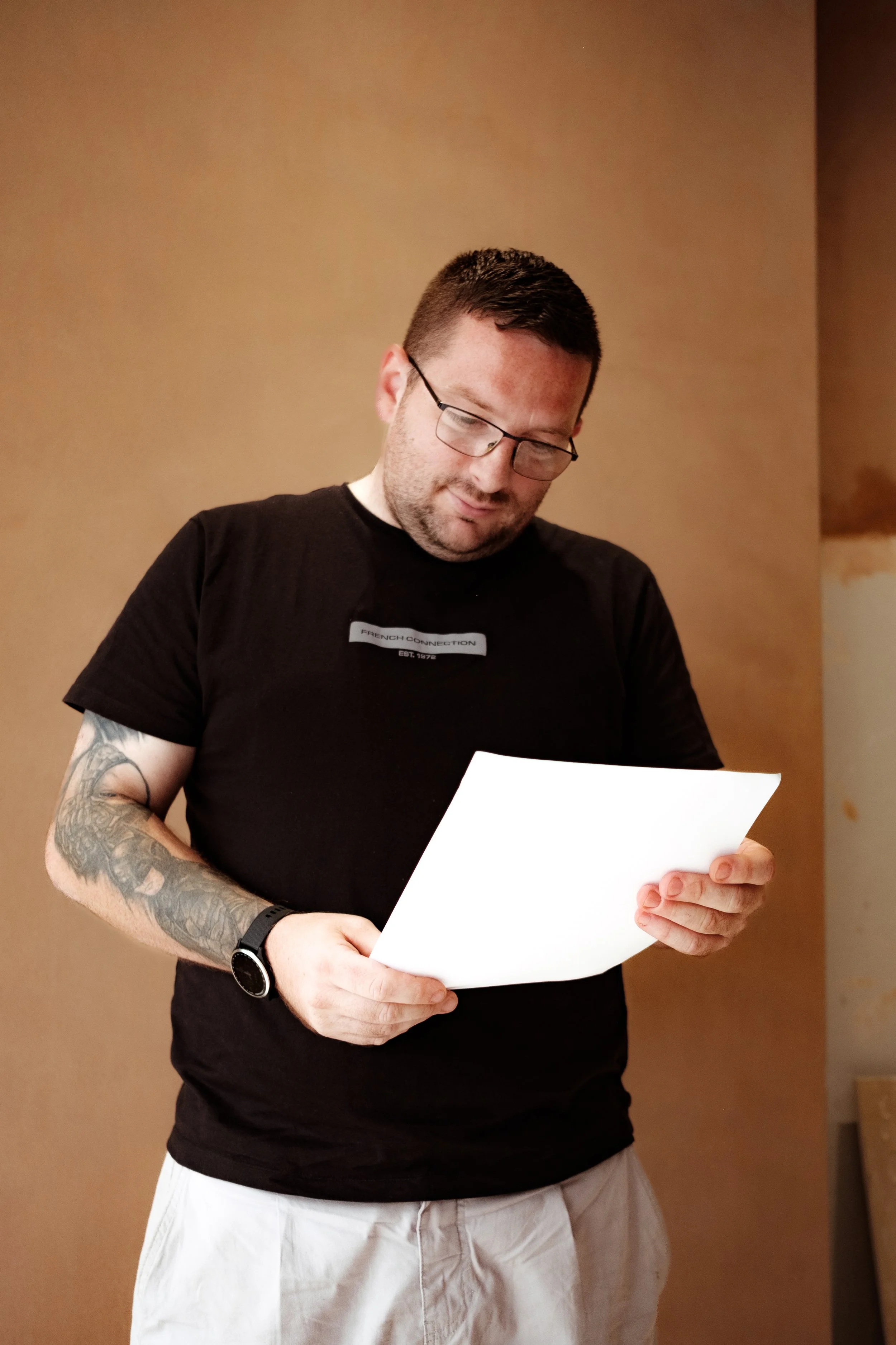 A man wearing glasses and a black T-shirt with a patch reading 'French Connection' is looking at a white sheet of paper he is holding in his hands. He has a tattooed arm and is wearing a black watch. He is standing indoors against a plain, light-colo