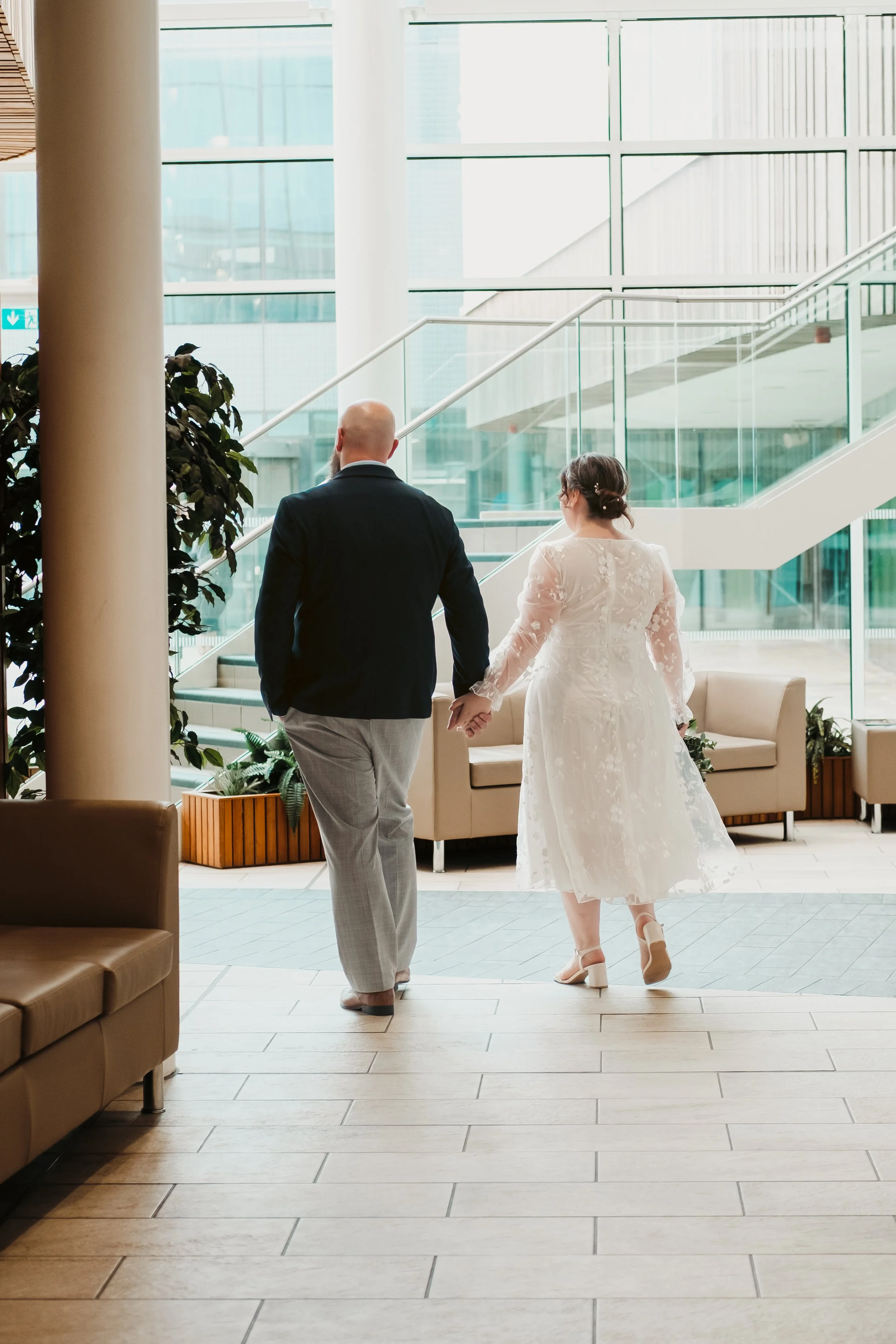 A couple holding hands walking inside a modern building with large glass windows, green plants, and comfortable seating.