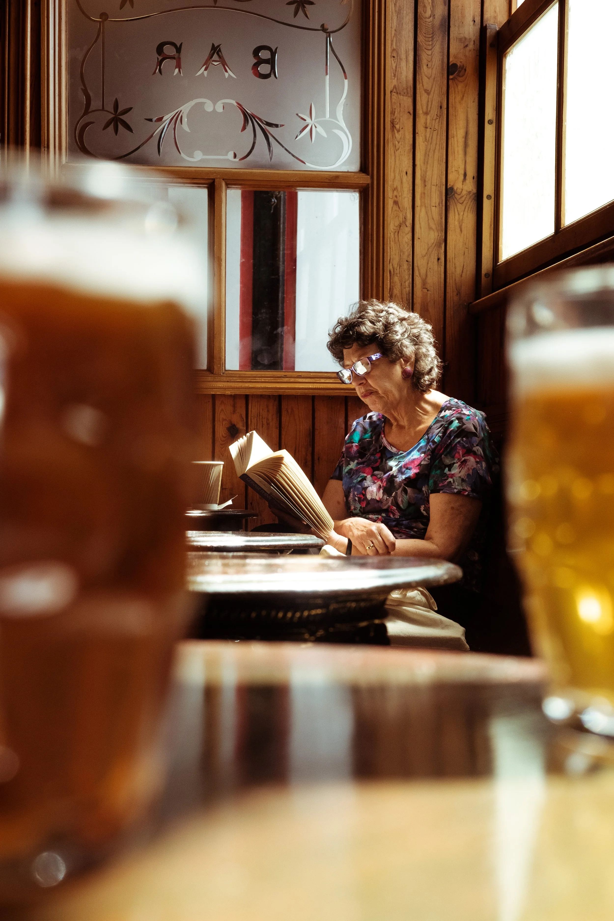 An elderly woman with curly hair and glasses reading a book in a rustic café with wooden walls and windows.