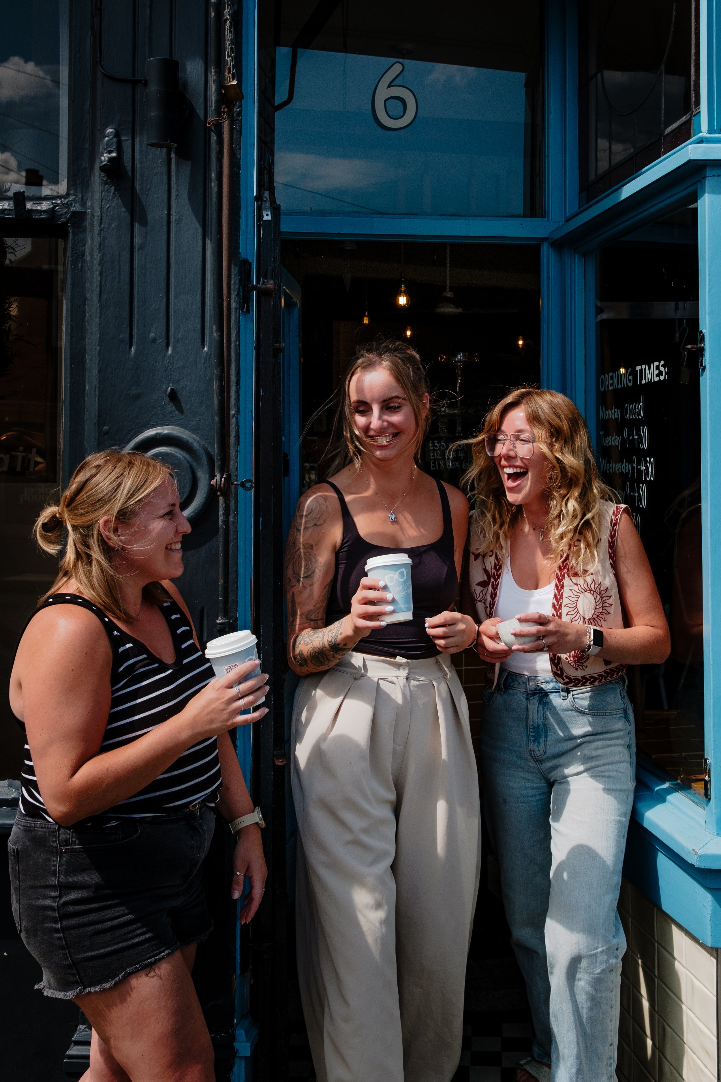 Three women standing outside a shop, smiling and holding coffee cups, engaging in conversation.