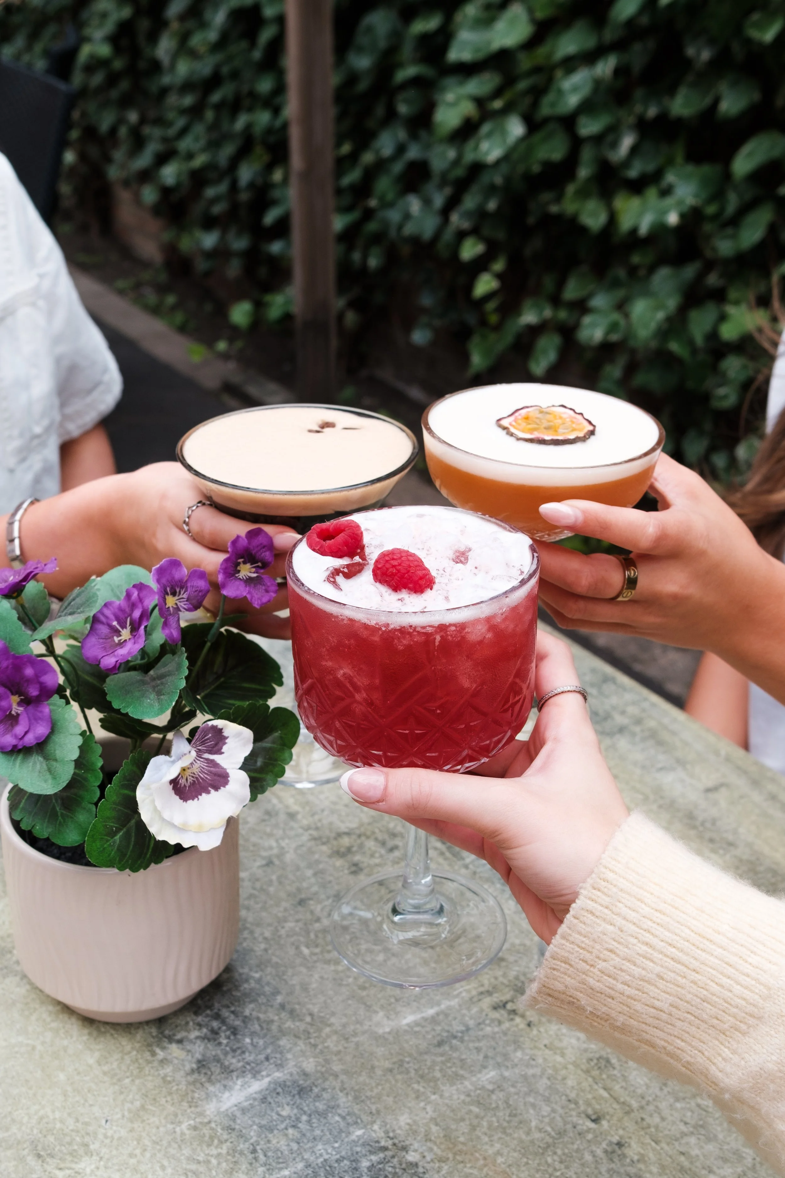 People holding cocktails with berries and passion fruit garnishes over a marble table, with purple flowers in a beige pot in front, and lush green foliage in the background.