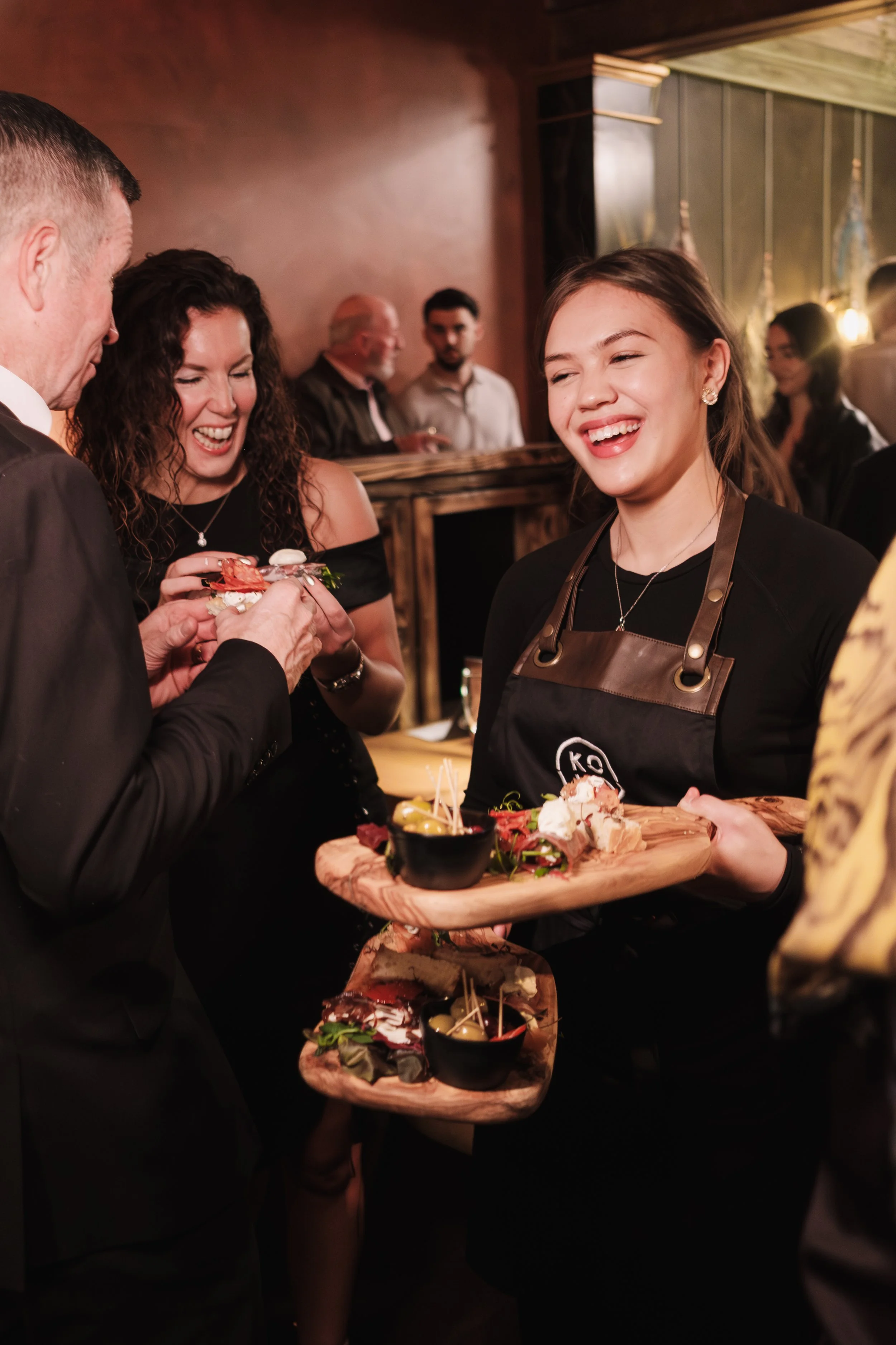 A young woman wearing a black apron serving food and drinks on a wooden tray at a social gathering or party, smiling and engaging with guests.