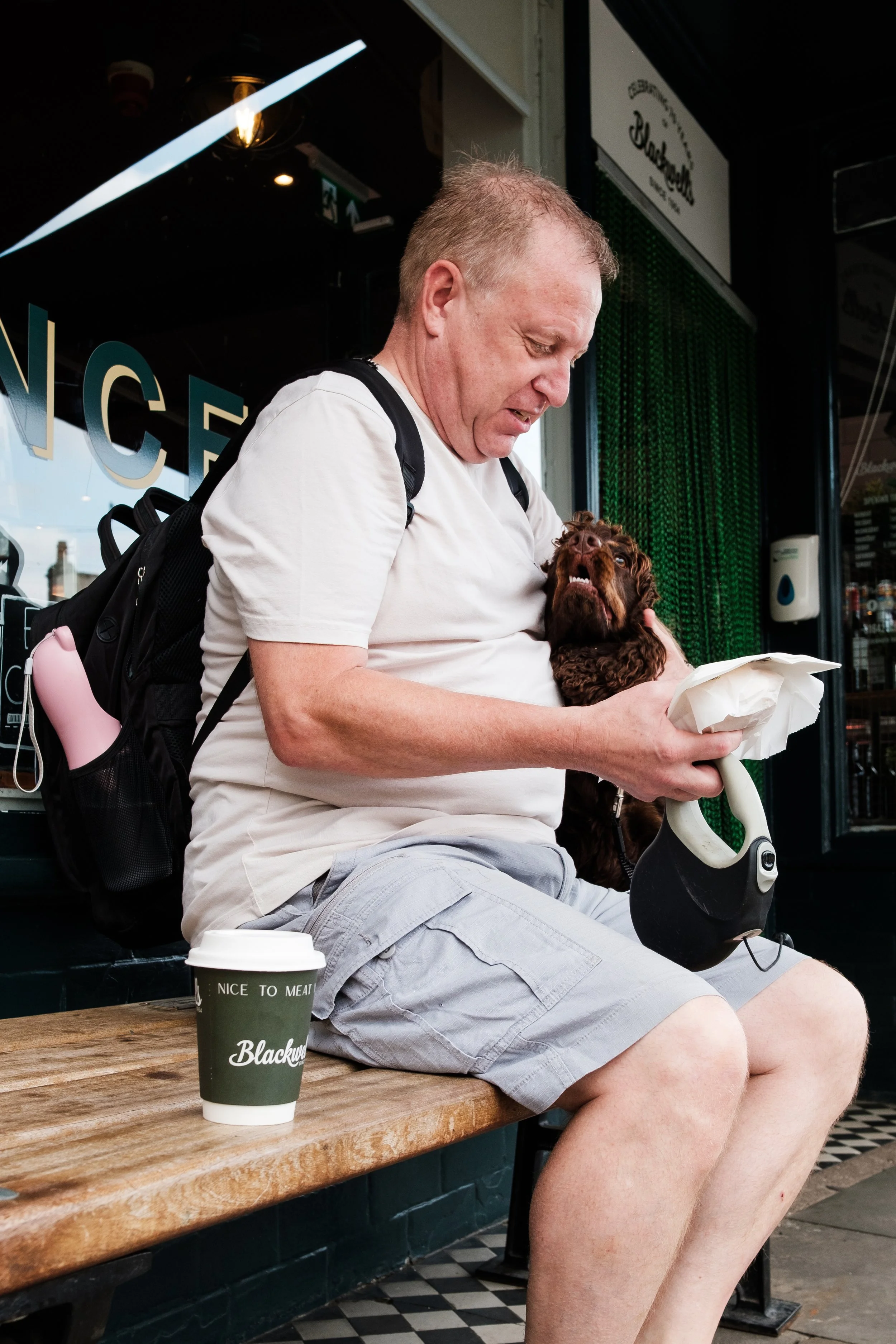 A man sitting on a bench outside a cafe holding a small brown dog in his lap, with a coffee cup on the bench nearby.