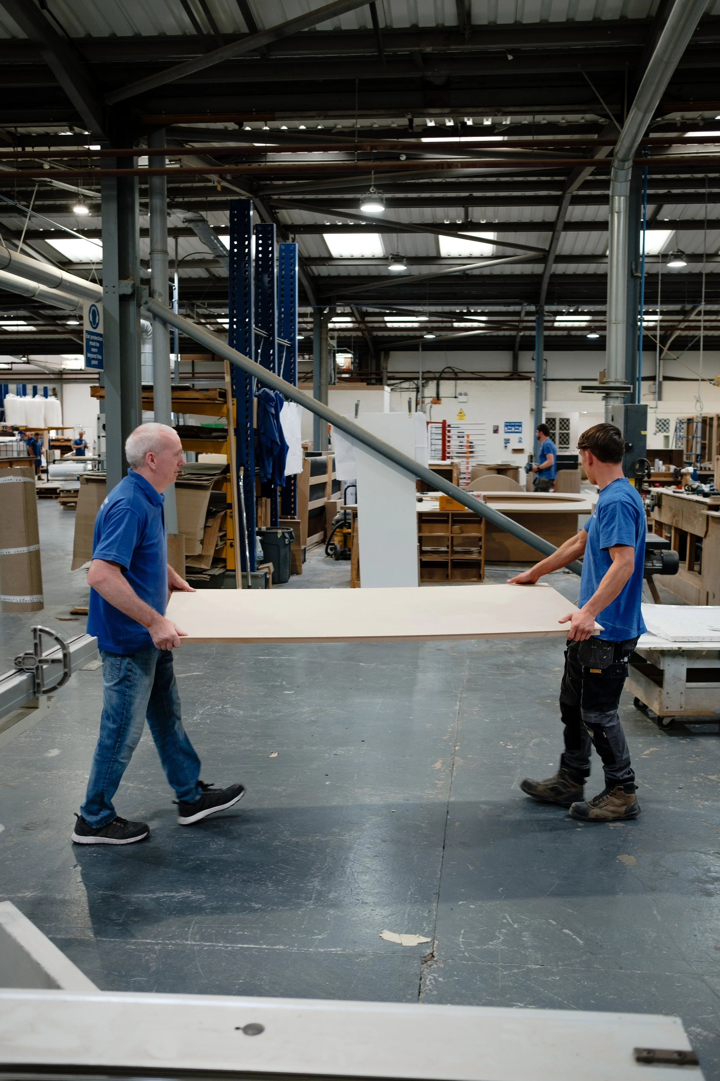 Two workers in blue shirts are passing a large sheet of wood in a woodworking shop. The shop has various tools, shelves, and other workers in the background.