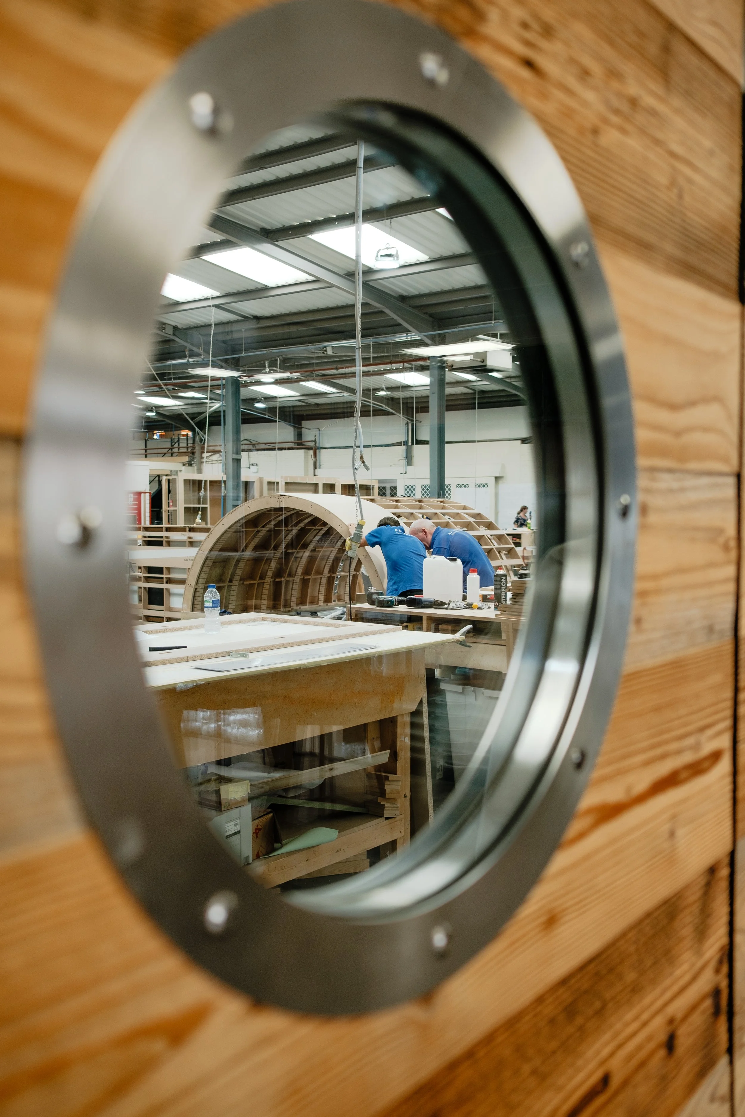 View through a circular window showing two people working on a wooden boat inside a workshop.