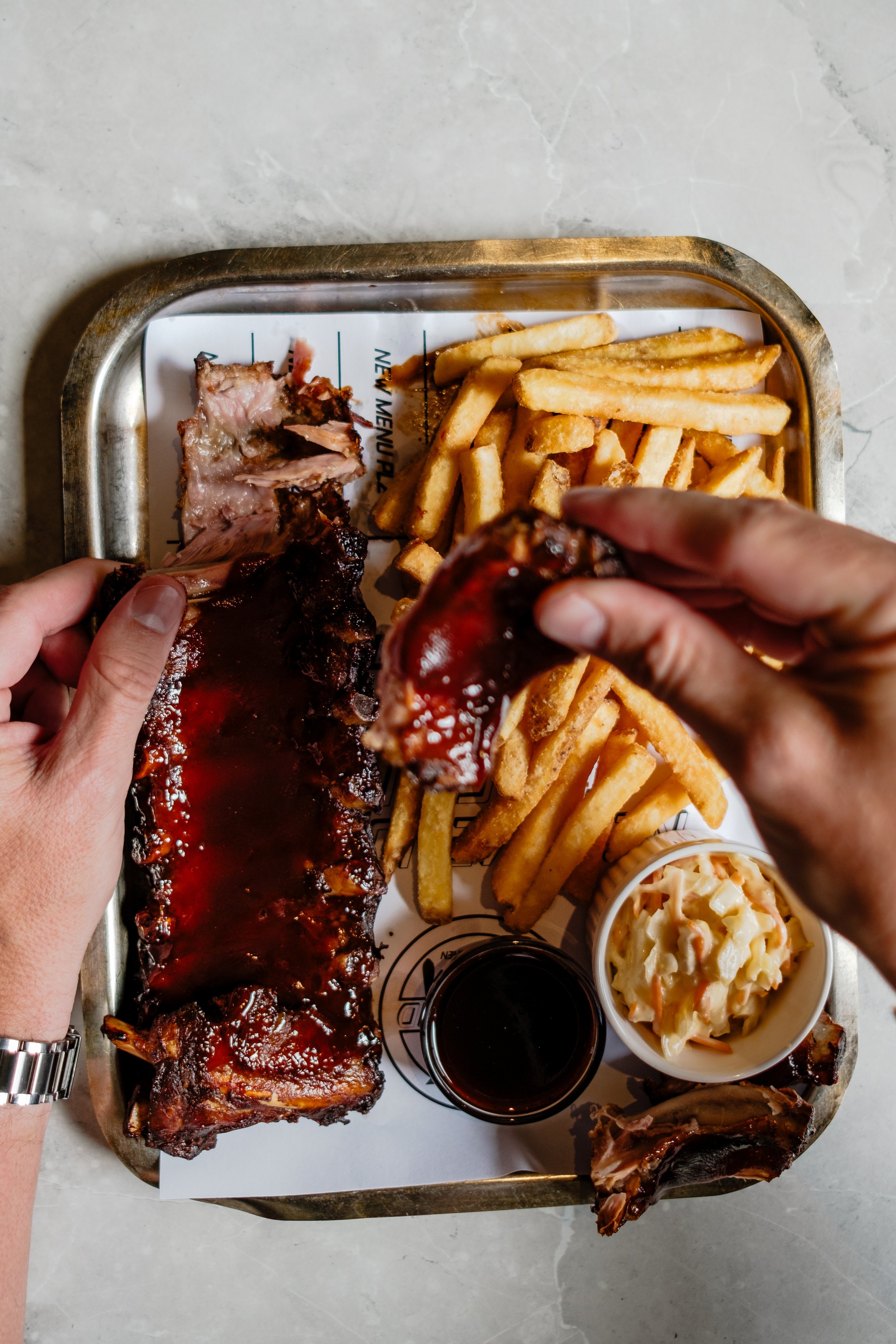 A tray with barbecue ribs, French fries, coleslaw, and a glass of dark soda, with hands holding a rib and a piece of meat.