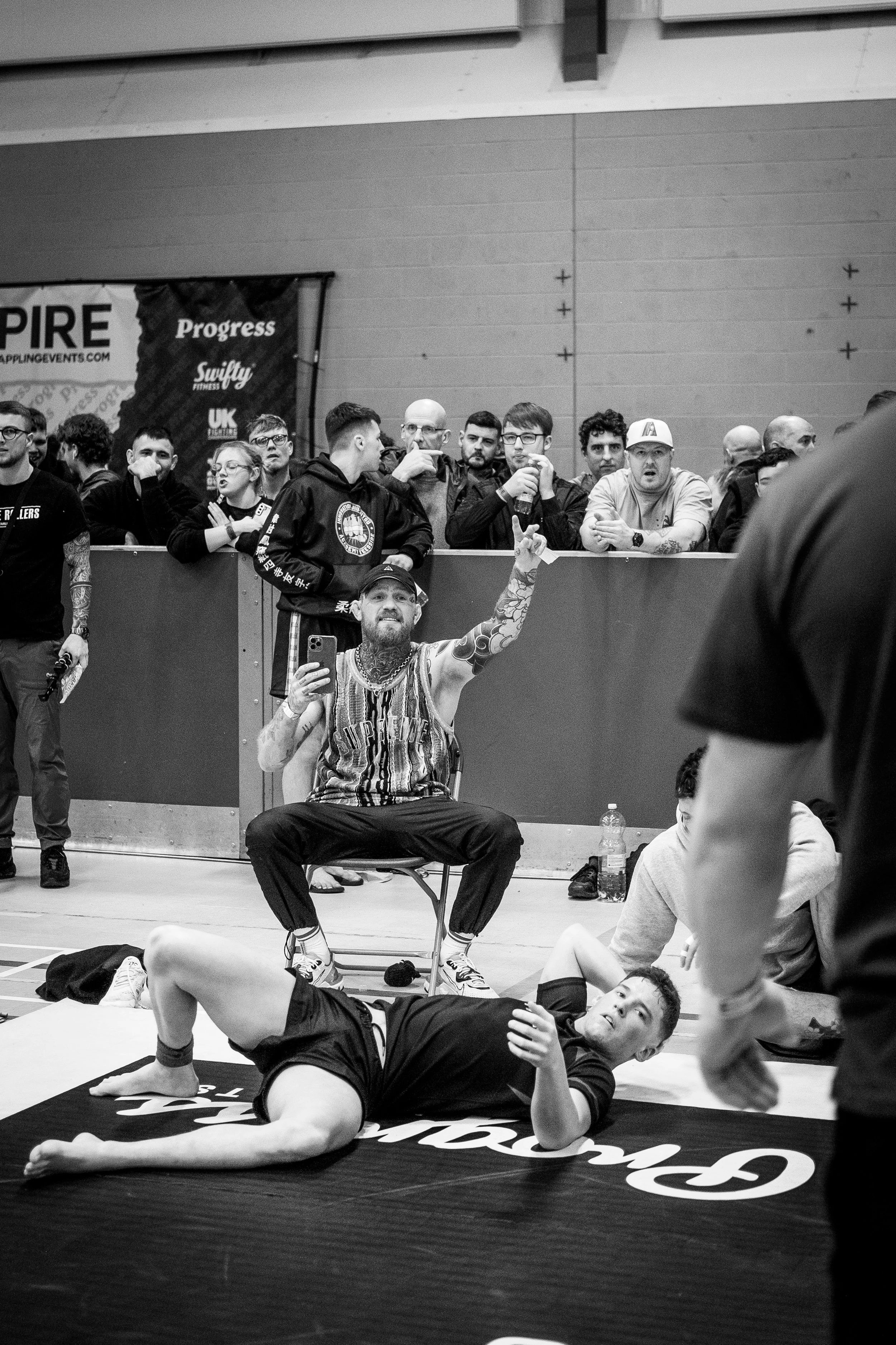 A man with tattoos taking a selfie while sitting on a chair behind a person lying on the ground at a fitness or sports event with a crowd of spectators observing.