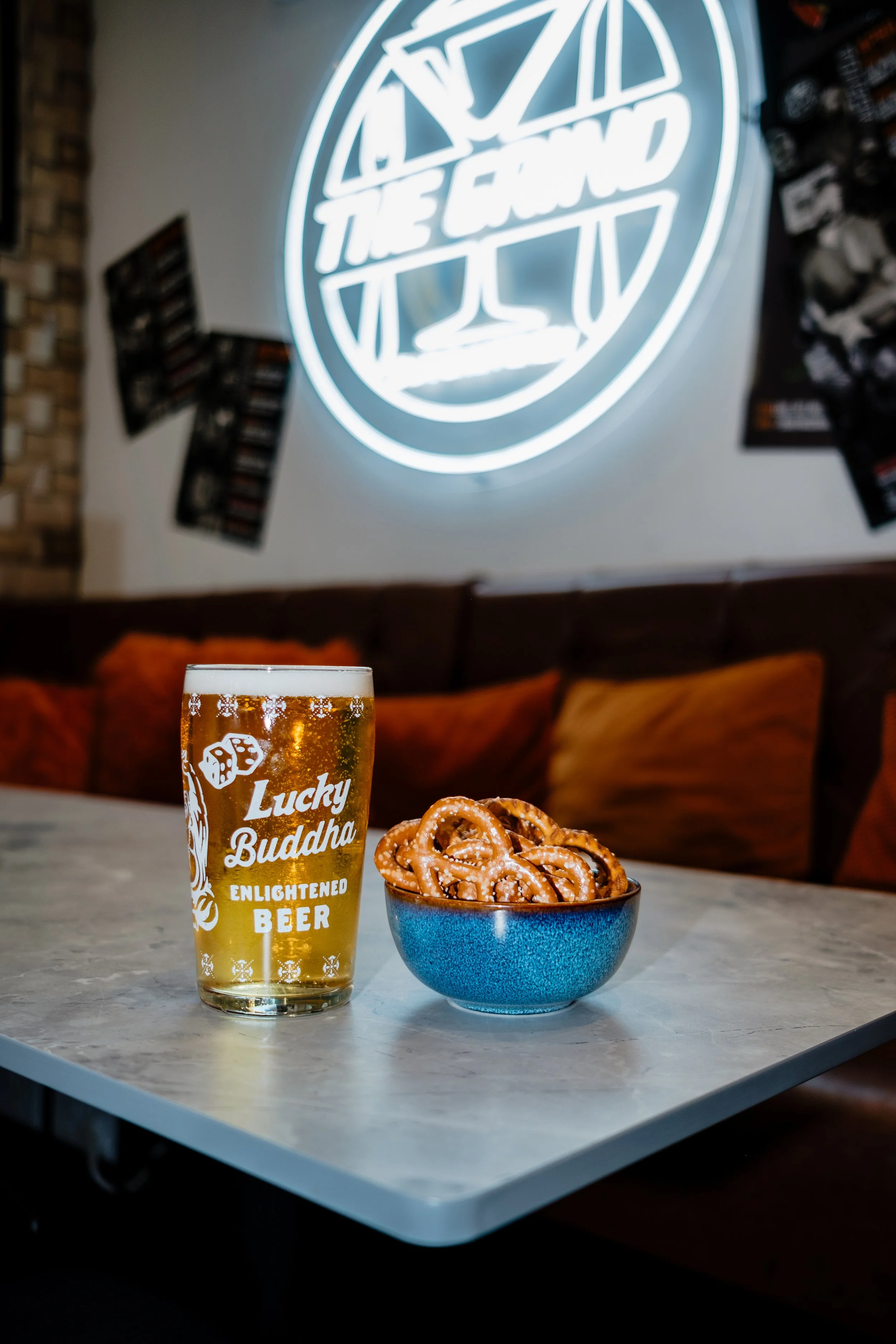 A glass of beer labeled Lucky Buddha Enlightened Beer and a bowl of pretzels on a marble table in a cozy bar or restaurant with a neon sign of a cocktail glass on the wall behind.