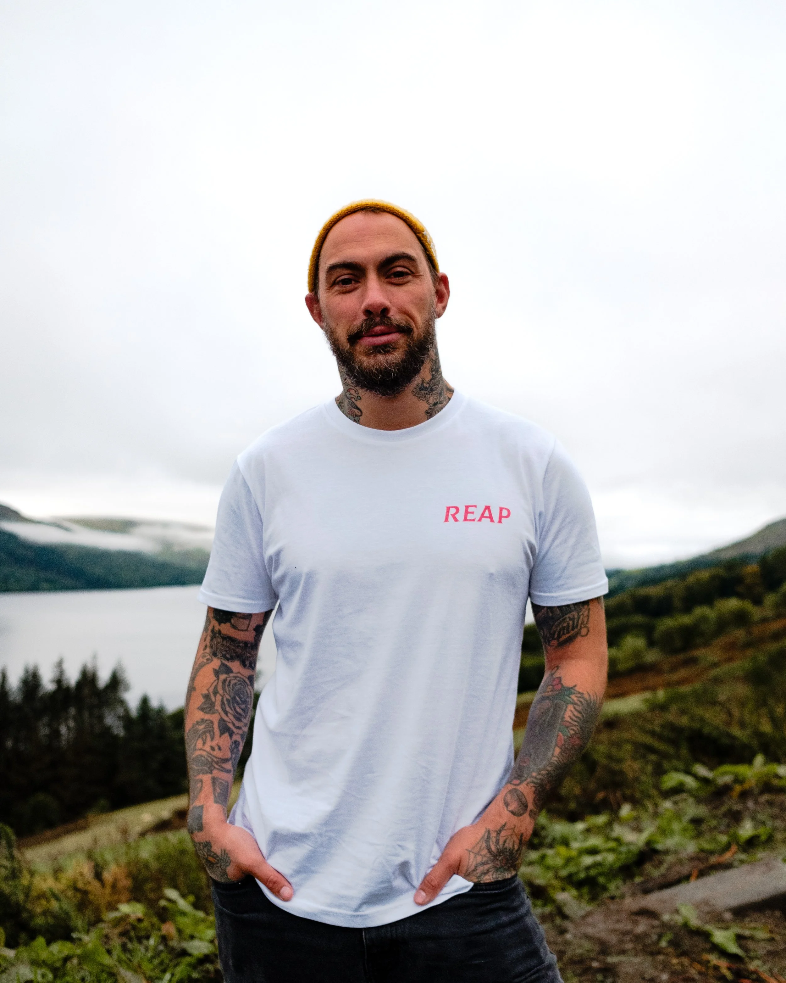 A man with a beard and tattoos on his arms and neck, wearing a yellow beanie and a white t-shirt with red text, stands outdoors with greenery and water in the background.