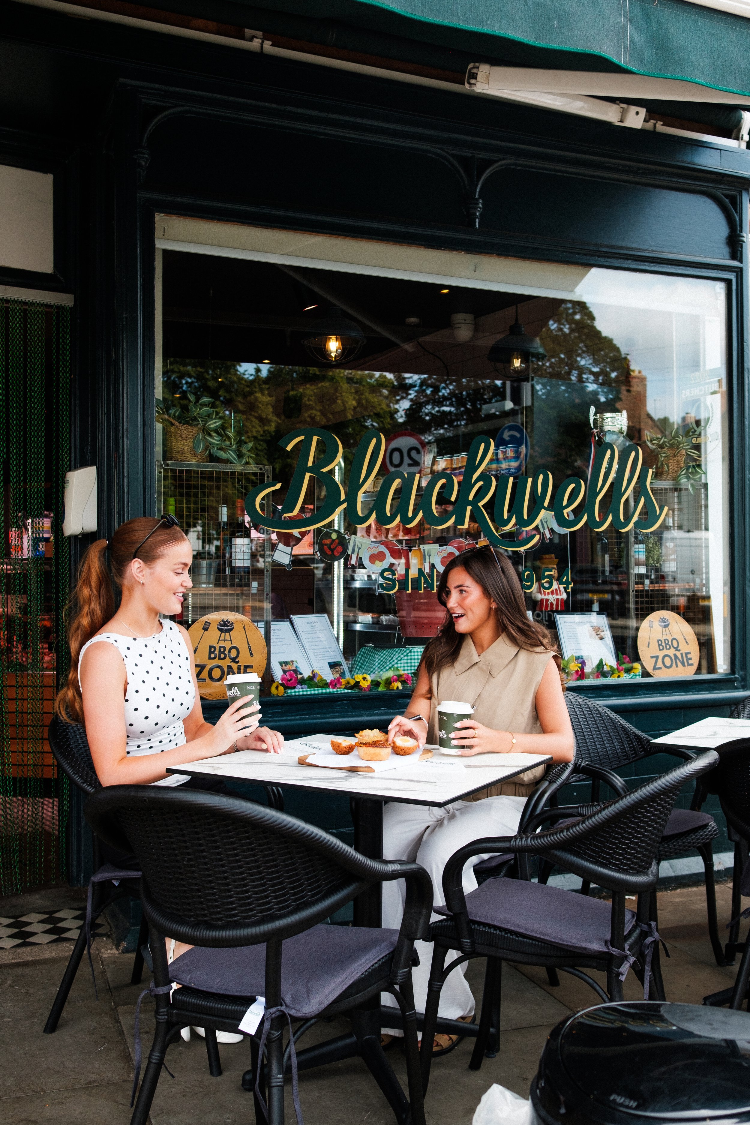 Two women sitting at an outdoor cafe table in front of a store window named Blackwells, enjoying coffee and pastries, with signs advertising a BBQ zone inside.