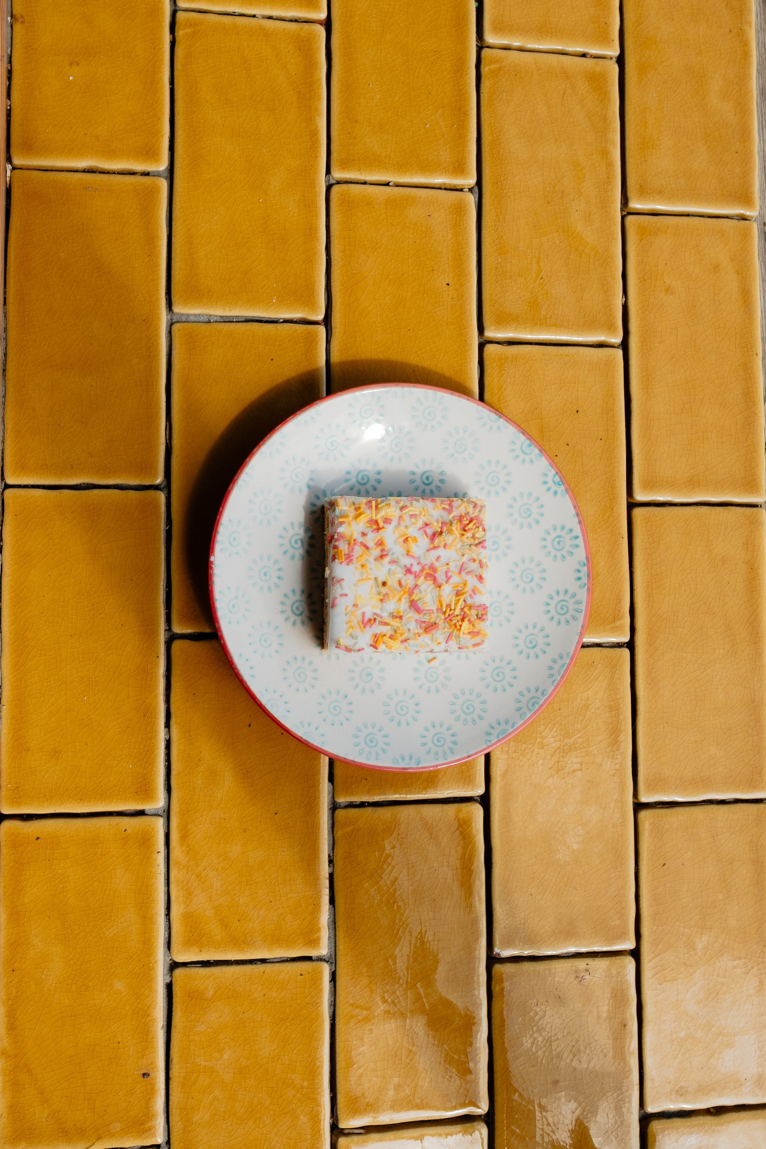 Top view of a plate with a slice of frosted cake topped with colorful sprinkles, placed on a tiled yellowish-brown surface.