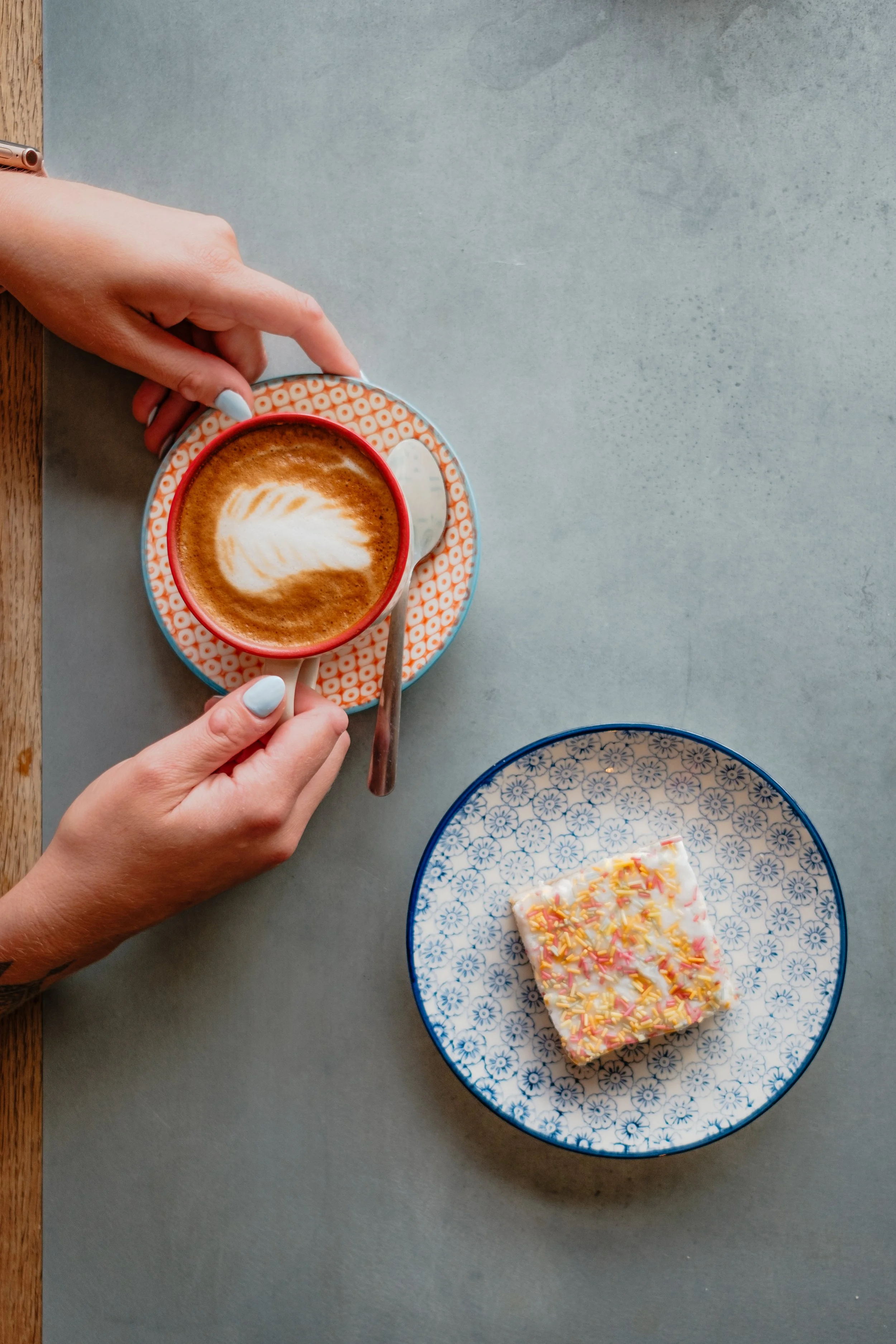 A person holding a cup of coffee with latte art, sitting at a table with a plate of decorated cake.