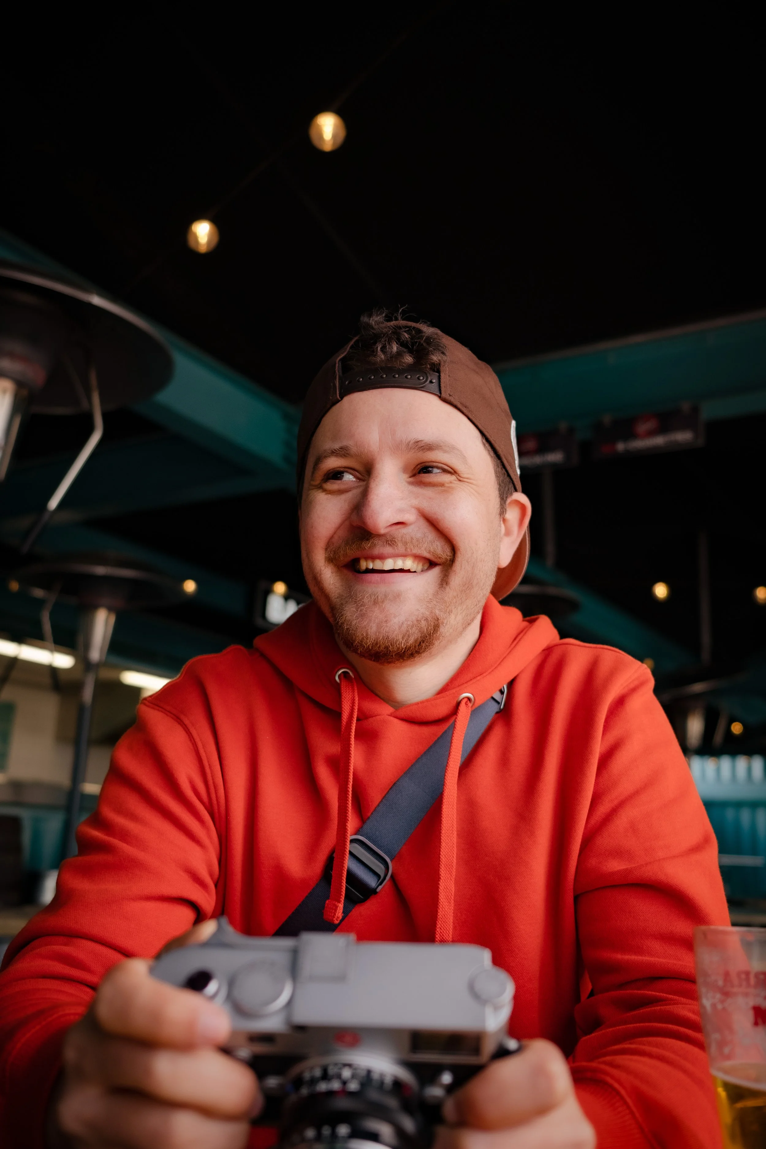 A young man with a beard and mustache wearing a backward brown cap and red hoodie, smiling and holding a video game controller.