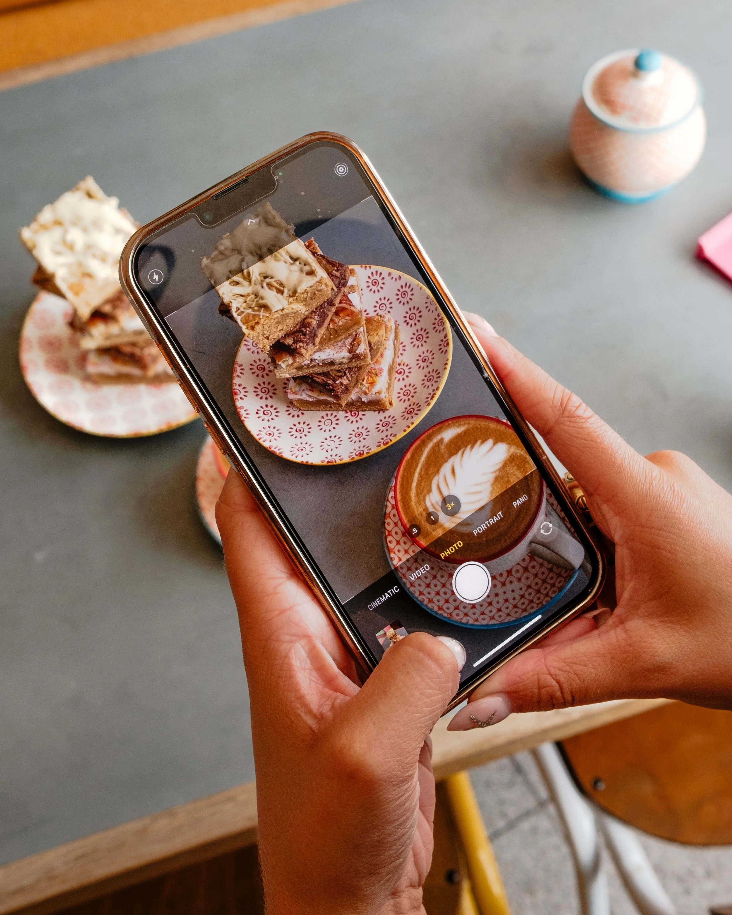 A person holding a smartphone taking a photo of plates of layered dessert bars and a latte on a gray table.