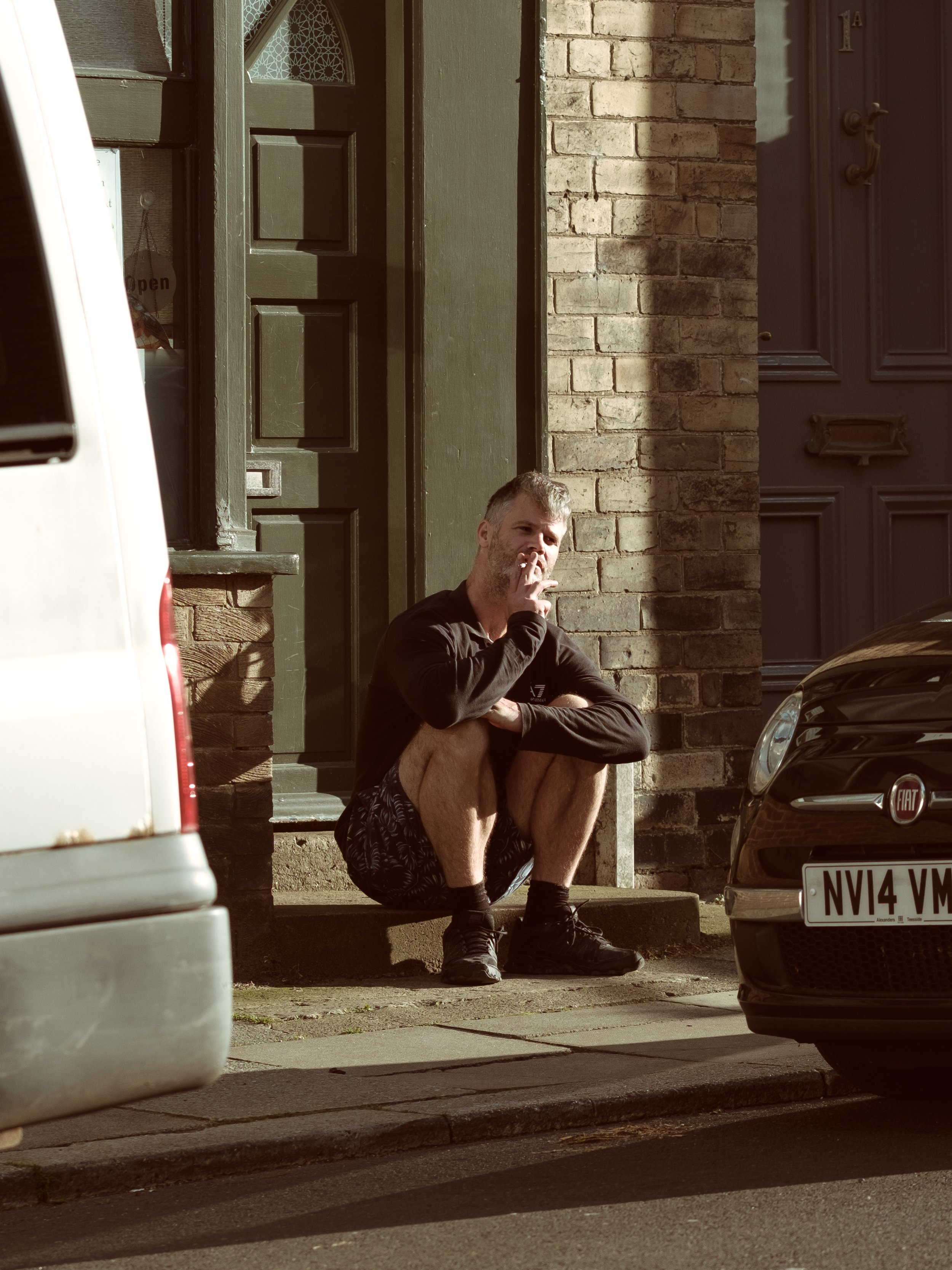 A man is squatting on the sidewalk in front of a brick building, smoking a cigarette. He is wearing a black long-sleeve shirt, patterned shorts, and running shoes. Cars are parked nearby, with the front of a gray car visible on the right and part of 
