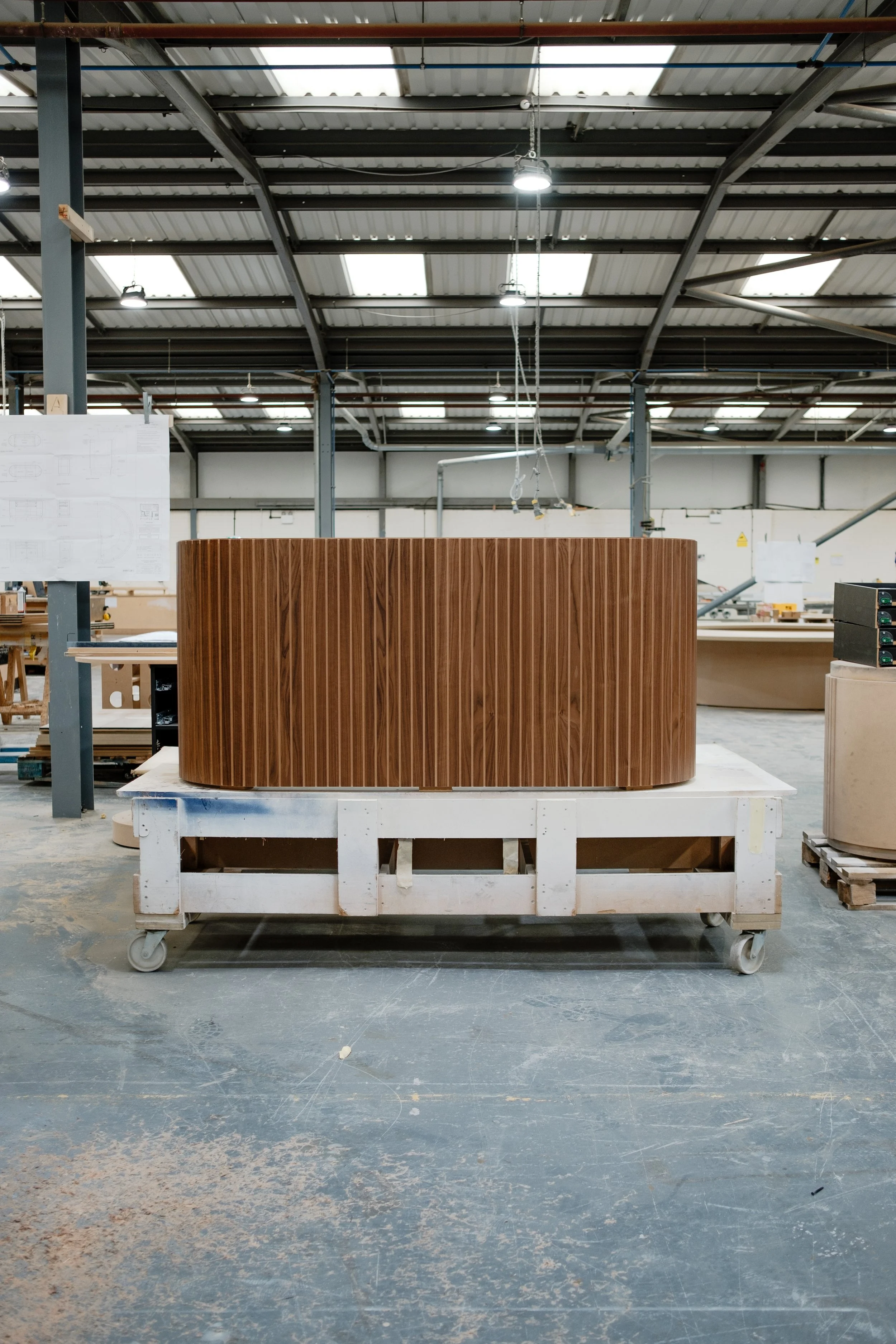 A large curved wooden structure, possibly a piece of furniture, placed on a white platform with wheels inside a spacious industrial workshop.