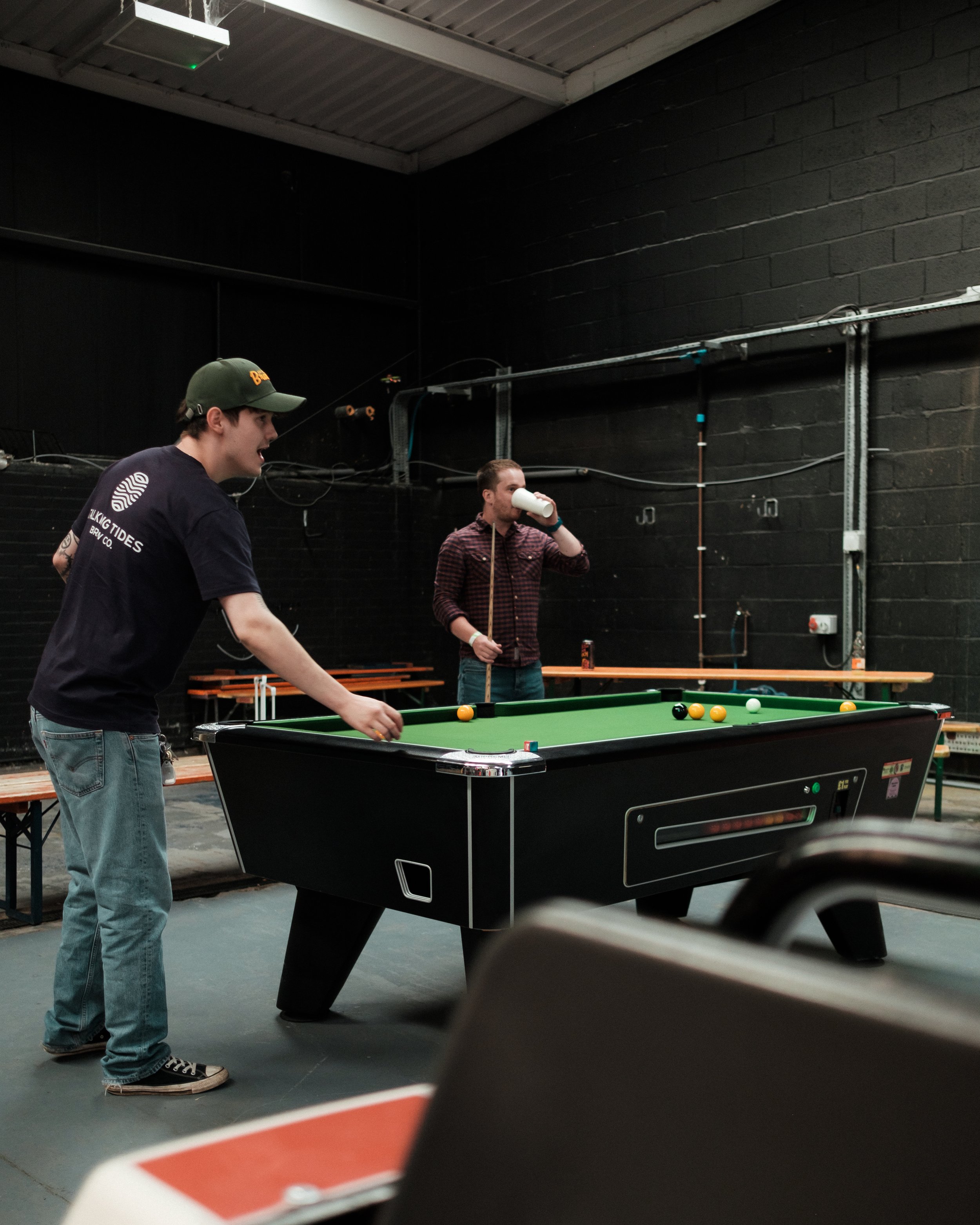 Two men playing pool in a game room with black walls and benches in the background.