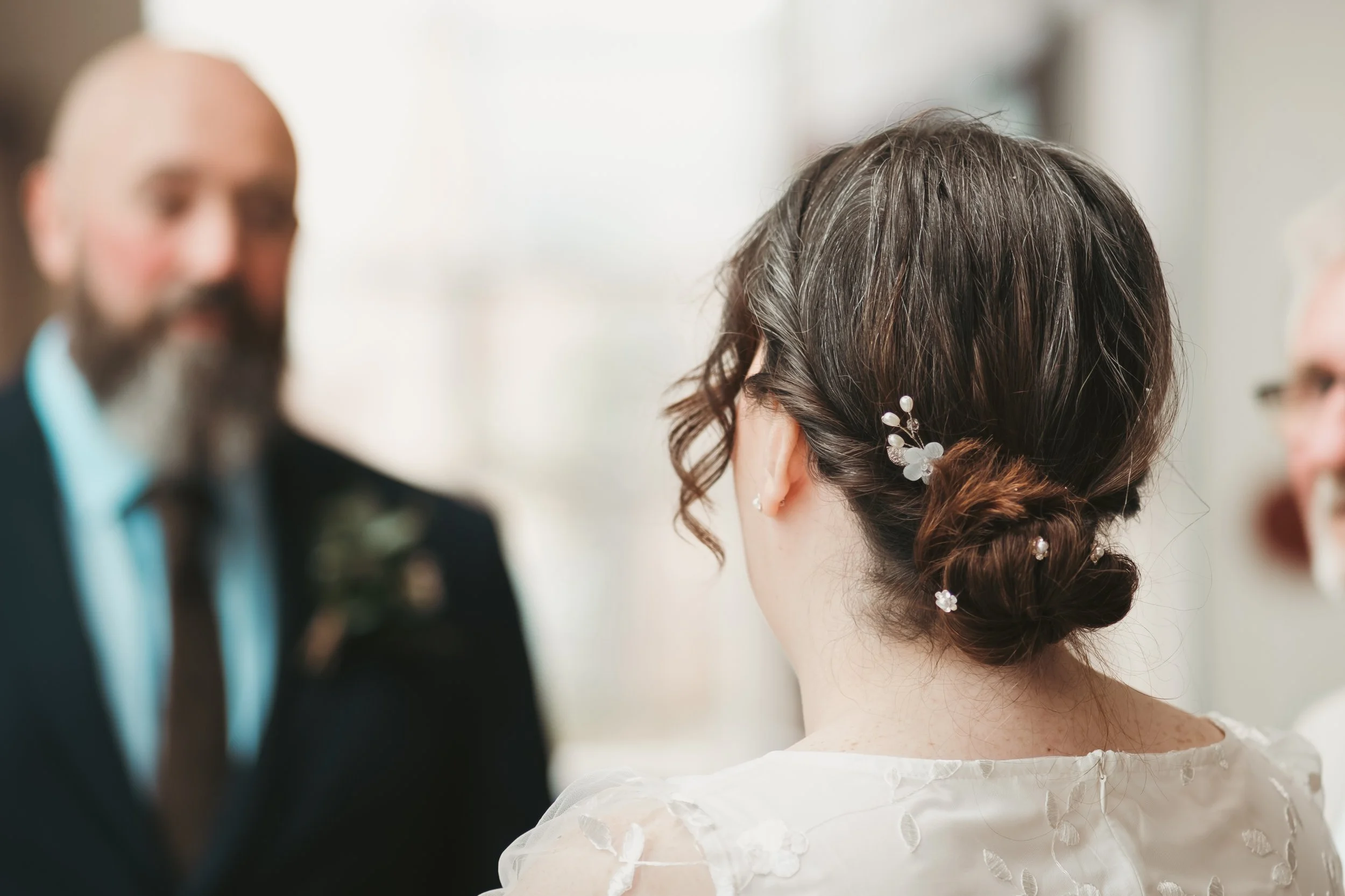 A woman with curly brown hair styled in an updo with white floral hair accessories faces a man with a beard, who is wearing a dark suit and light blue shirt, in a blurred background.