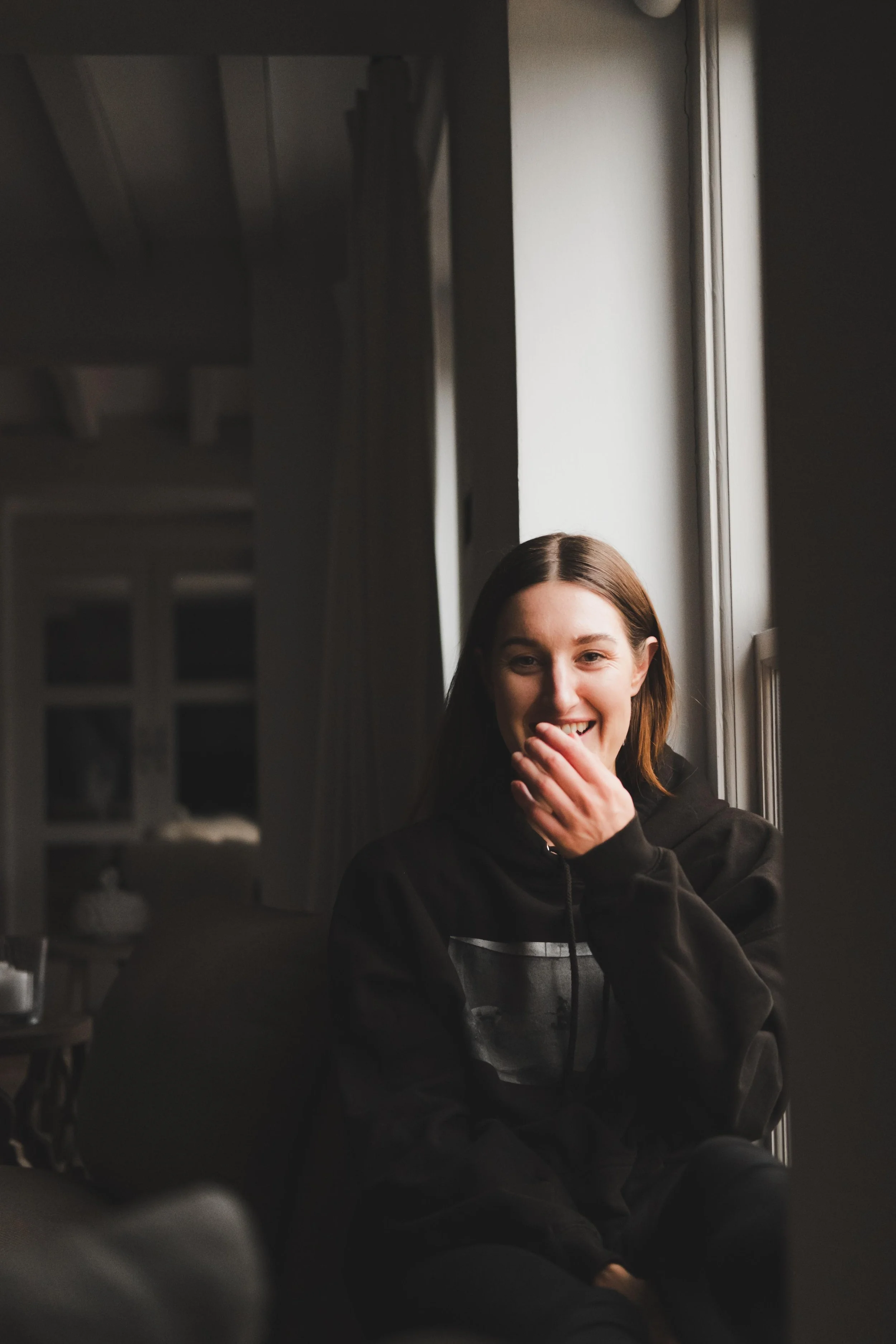 A young woman with brown hair and a big smile, sitting by a window, covering her mouth with her hand, wearing a black hoodie, in a dimly lit indoor setting.