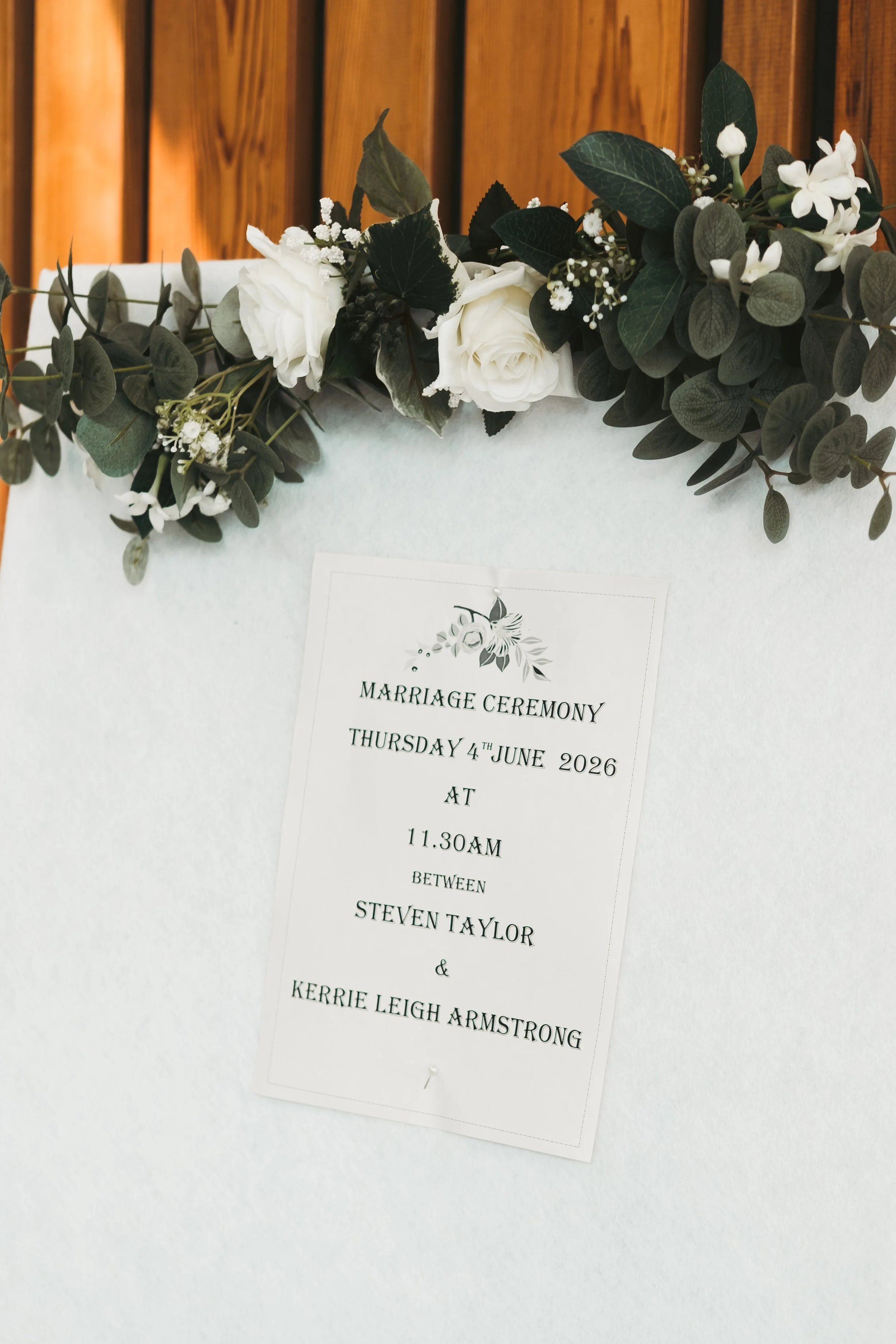 A floral decoration with white roses, dark green leaves, and small white filler flowers, placed above a wedding invitation on a white surface.
