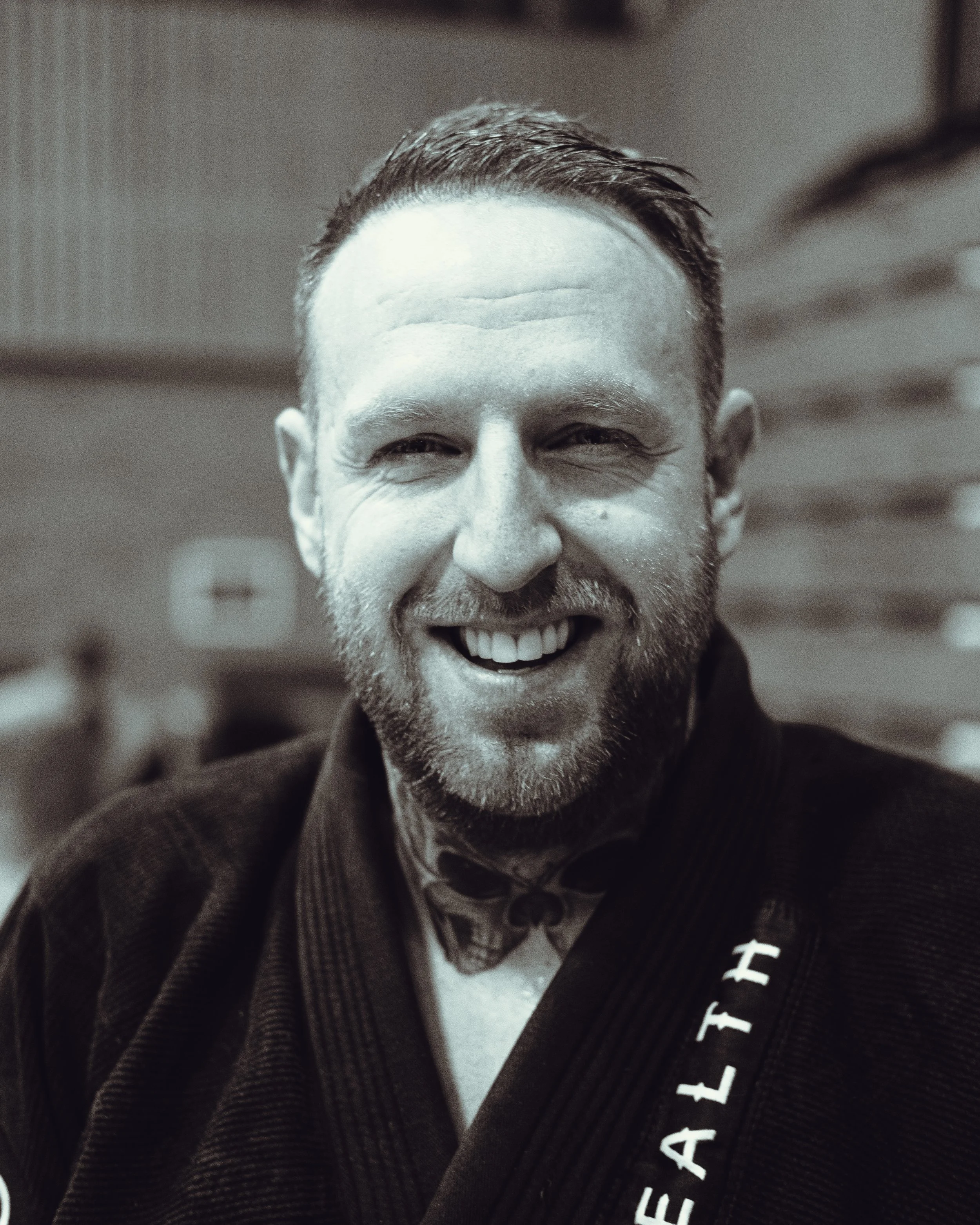 Close-up of smiling man with short hair, beard, and dark gi with white and red lettering.
