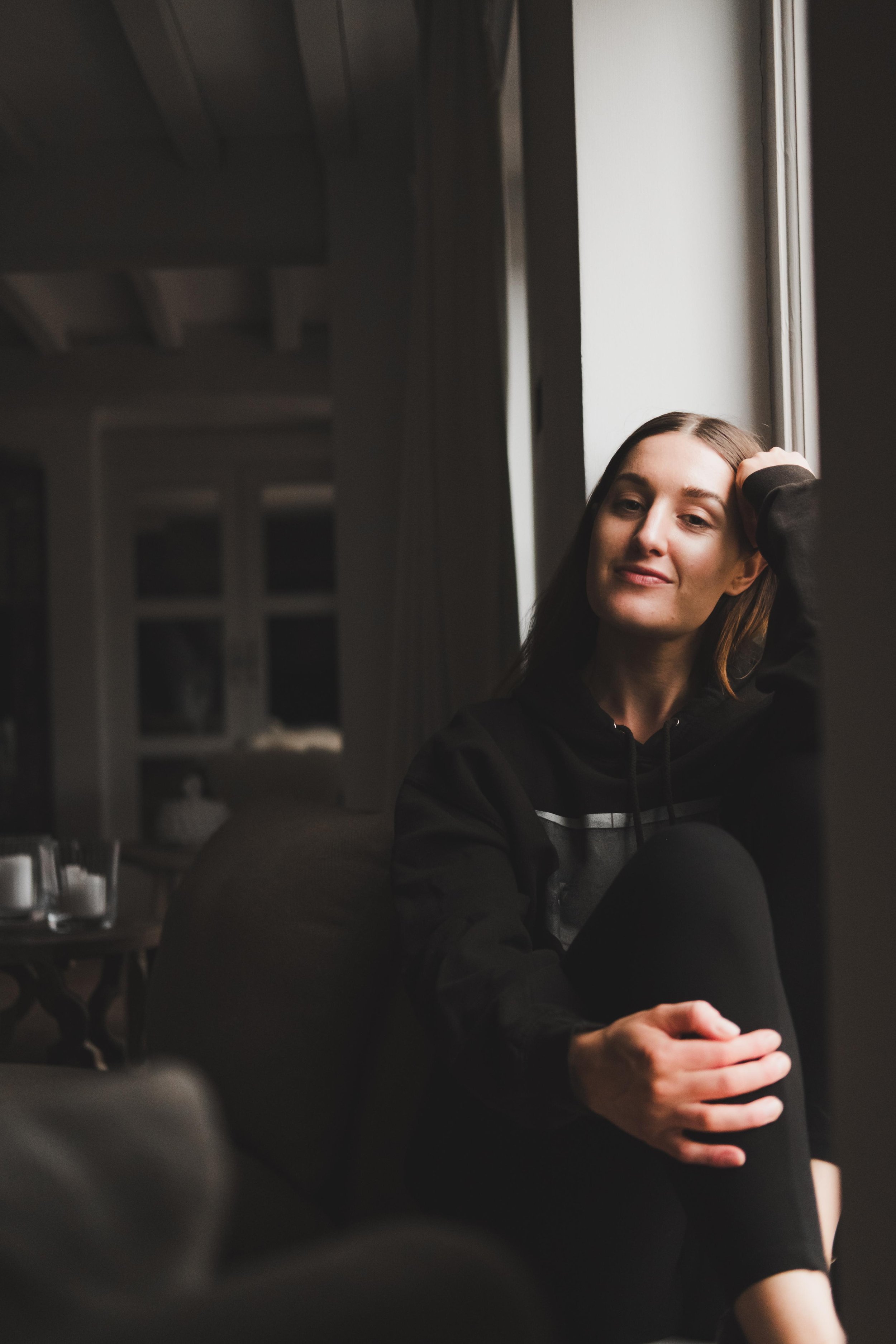 A young woman with long brown hair, wearing a black hoodie, sitting by a window with her hand on her head, smiling slightly, in a dimly lit indoor setting.