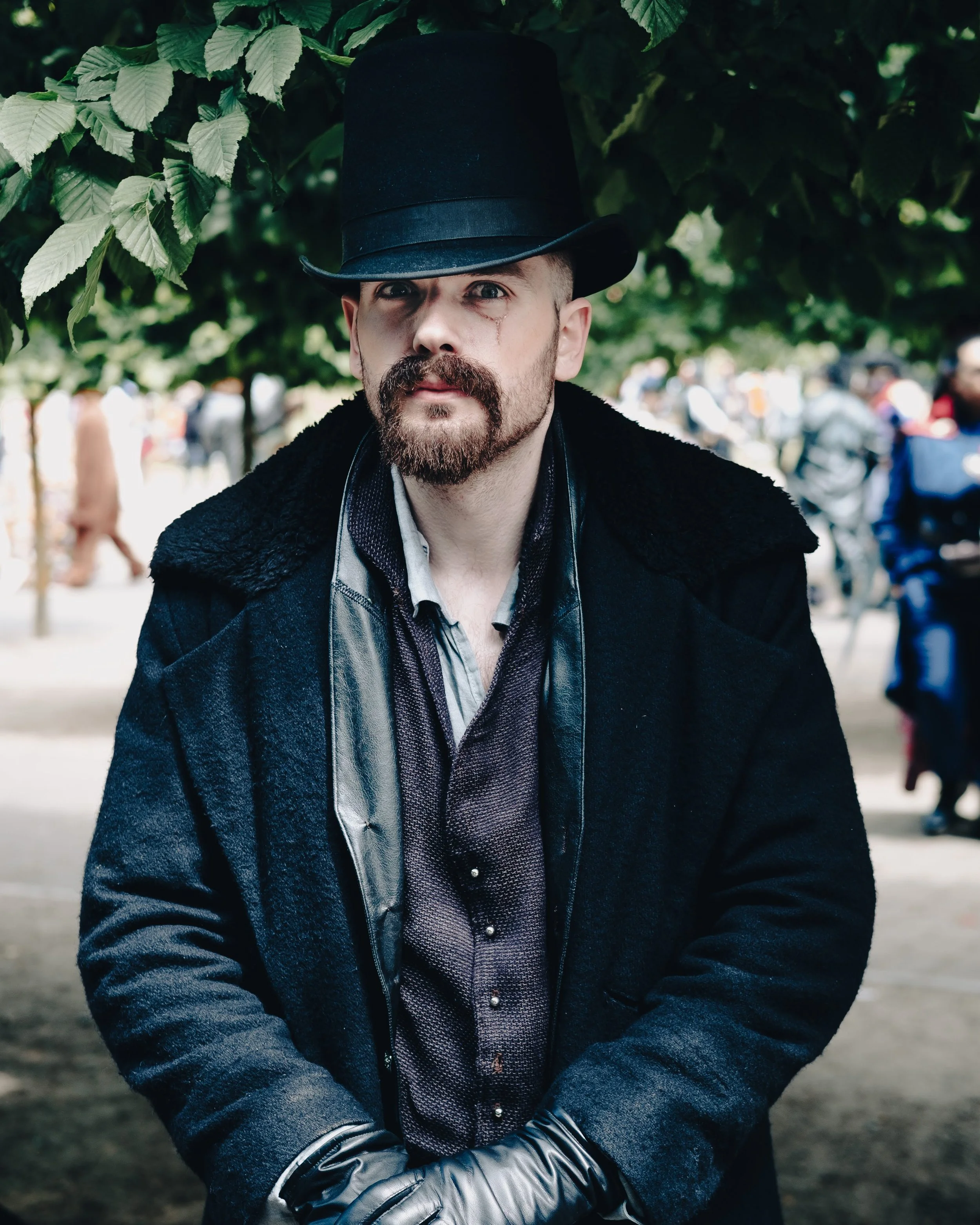 A man with a beard and mustache wearing a black top hat, black coat, and leather gloves standing outdoors in a park or garden with green leaves and other people in the background.