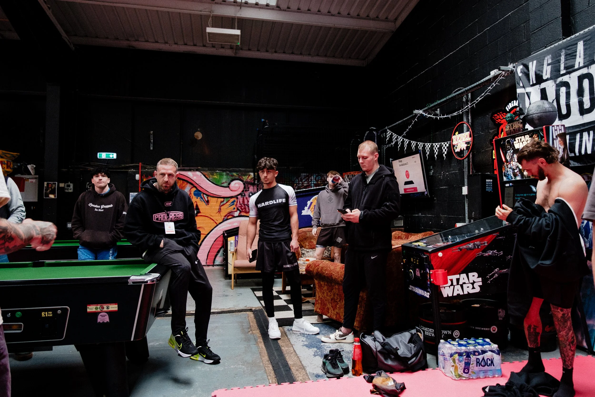 Group of young men gathered around a pool table in a dimly lit indoor space with graffiti on the walls, arcade machines, and Star Wars-themed decorations.