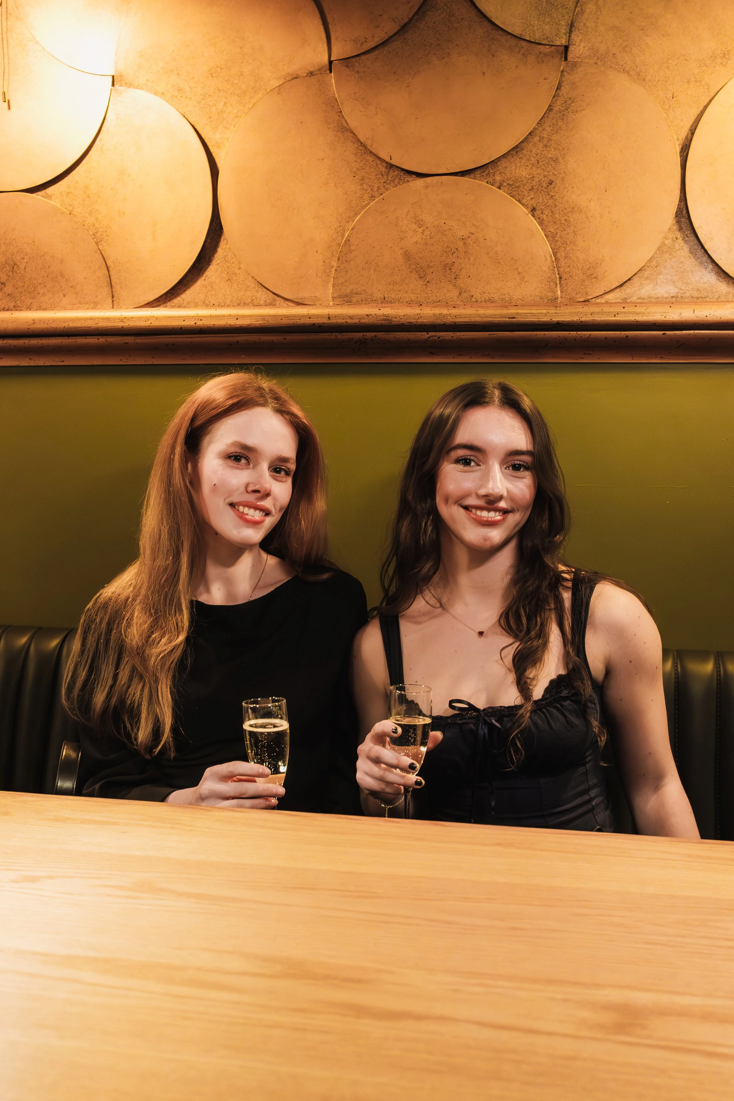 Two young women sitting at a bar or restaurant, smiling and holding glasses of champagne.