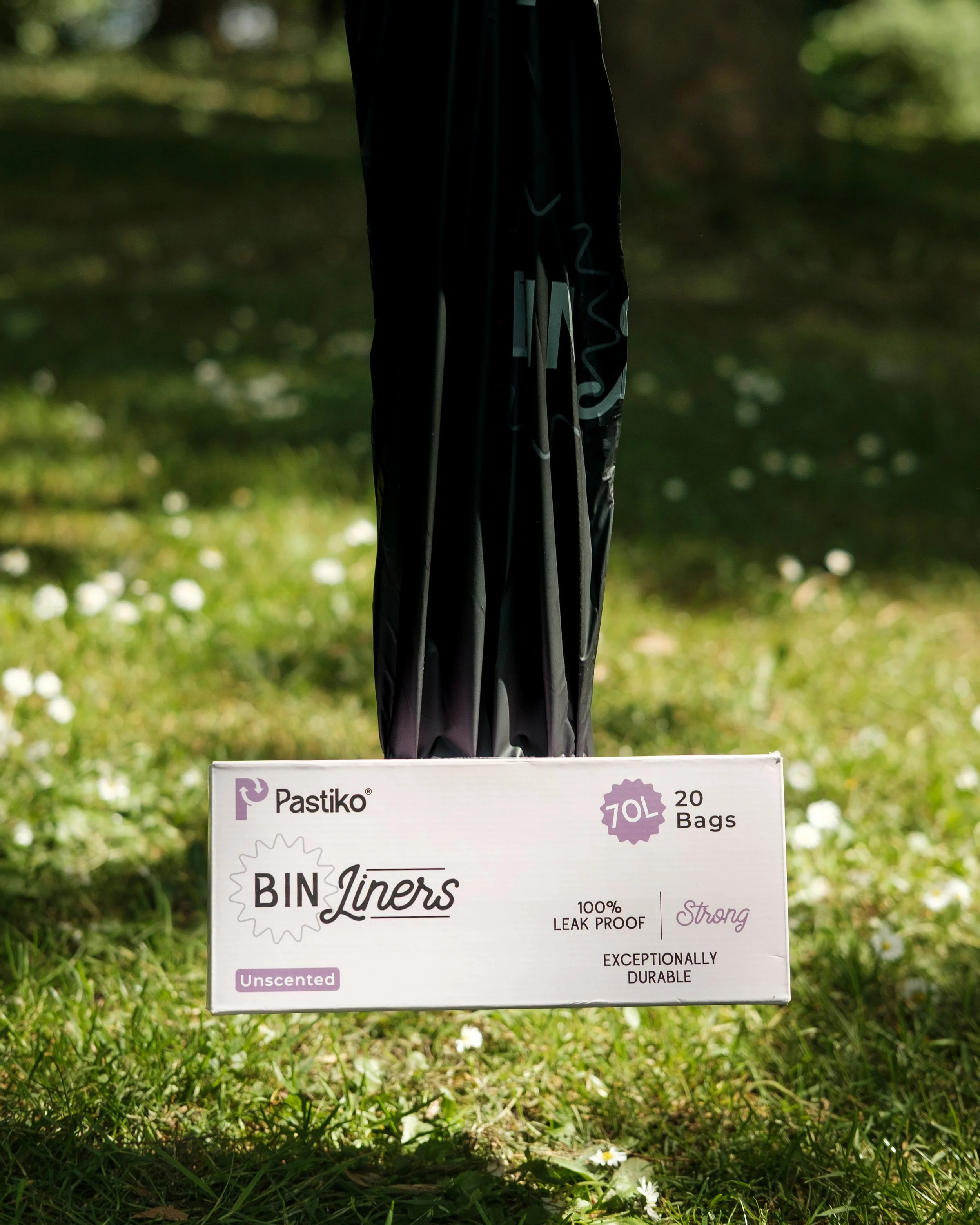 A box of Pastiko bin liners with a black trash bag hanging above it, set outdoors on green grass with white flowers, in front of a blurry natural background.