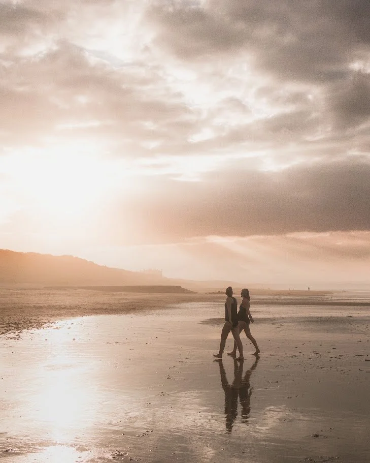 Two people walking along the beach during sunset with their reflections visible on the wet sand.