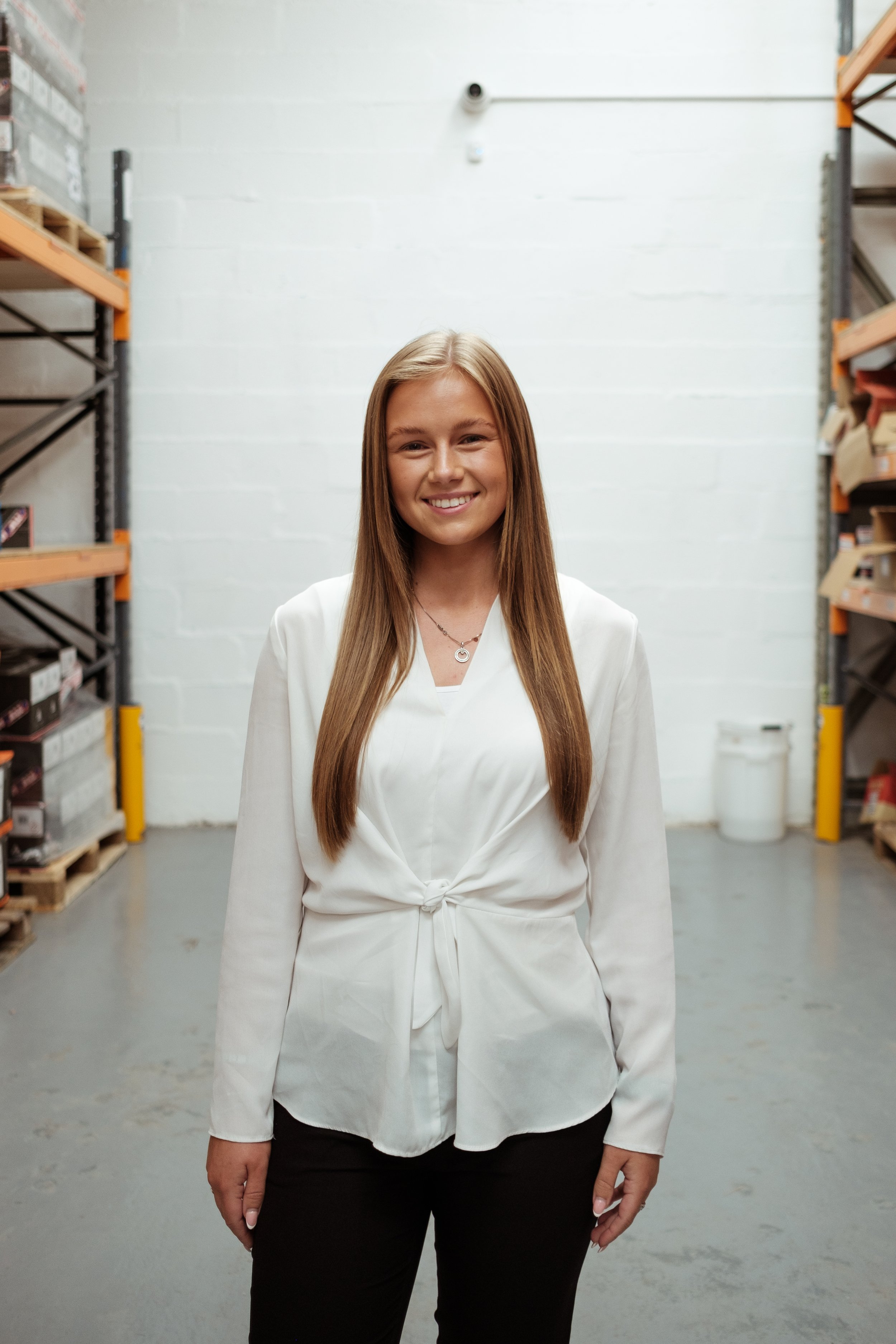 A young woman with long brown hair wearing a white blouse and black pants standing in a warehouse aisle with metal shelves on both sides filled with boxes and a white wall in the background.