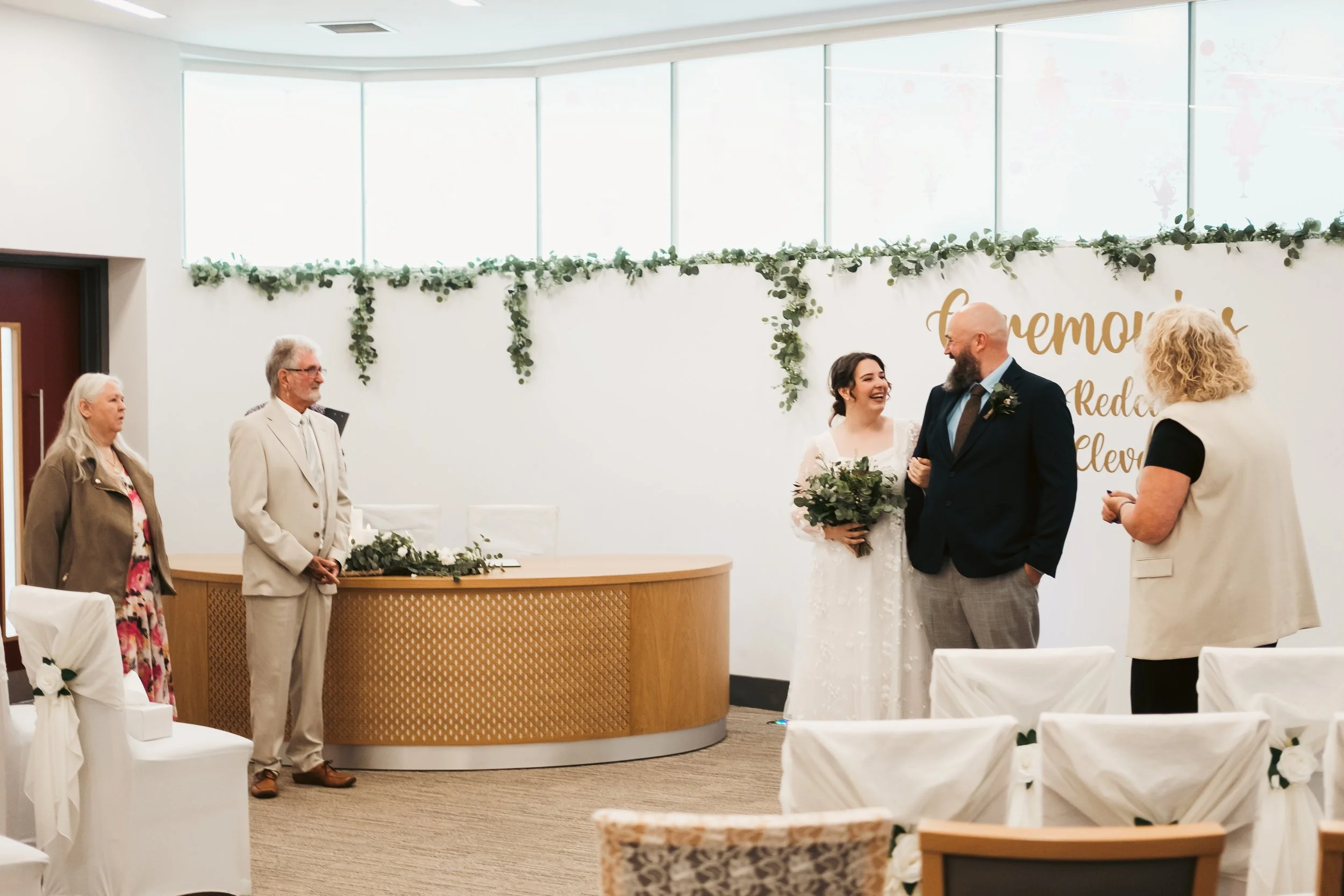 A wedding ceremony taking place in an indoor venue with white draped chairs and floral decorations. The bride and groom are smiling and holding hands, with a few guests standing nearby. Large windows with natural light and a backdrop with the word 'r