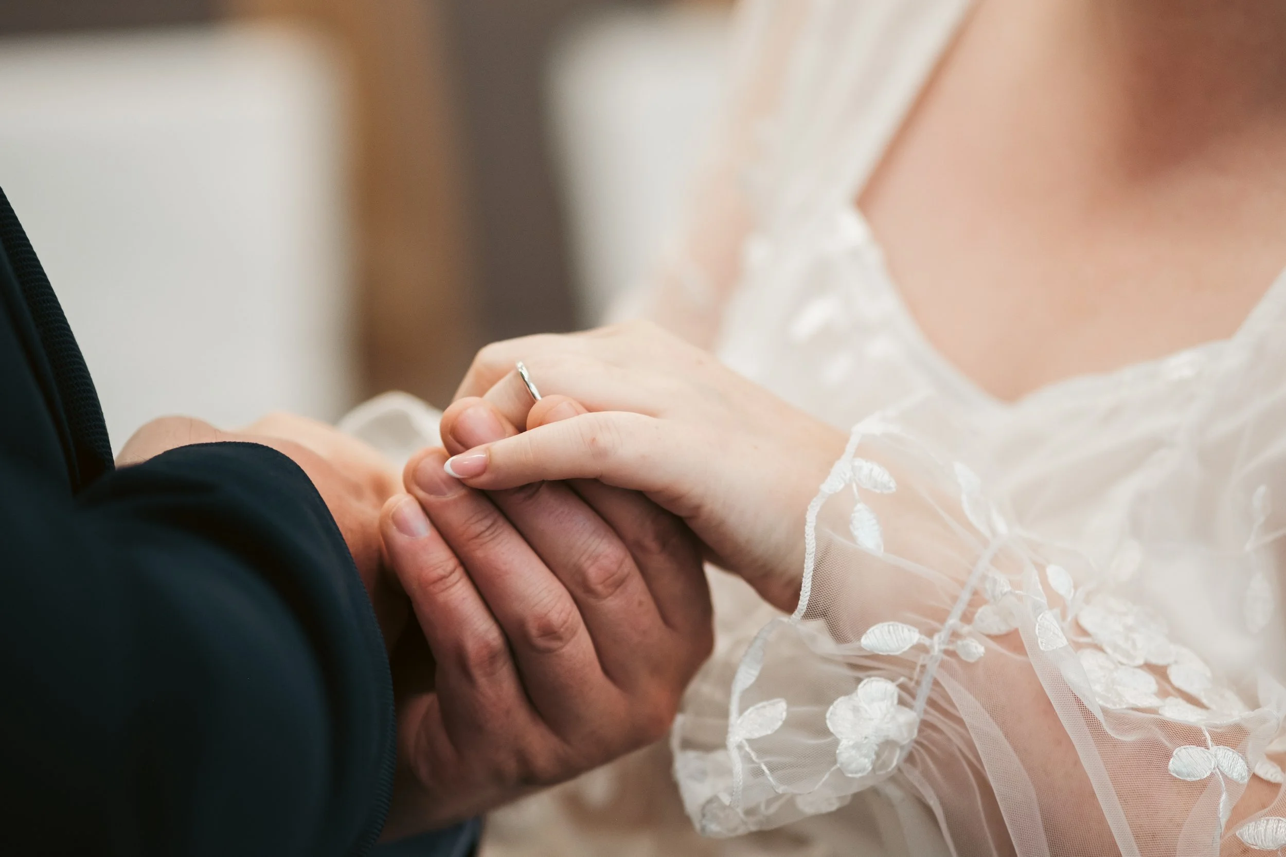 A close-up of a bride and groom holding hands during a wedding ceremony, with the bride wearing a white lace dress and the groom in a dark suit.