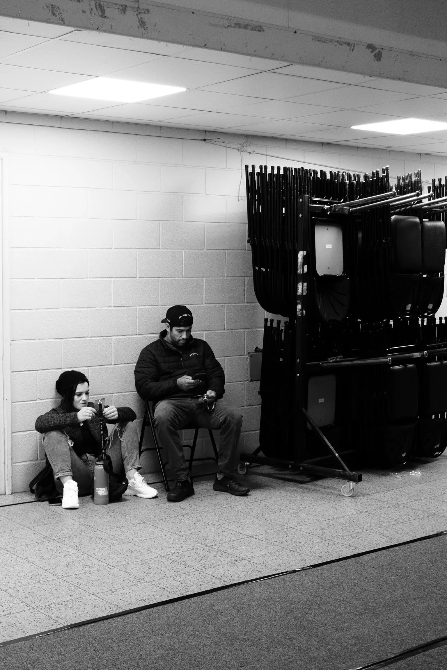 A black-and-white photo of two people sitting against a cinder block wall, both looking at their phones. One person, a woman, is wearing a beanie and sitting on the floor with a water bottle, while the other, a man, is wearing a cap and sitting on a 