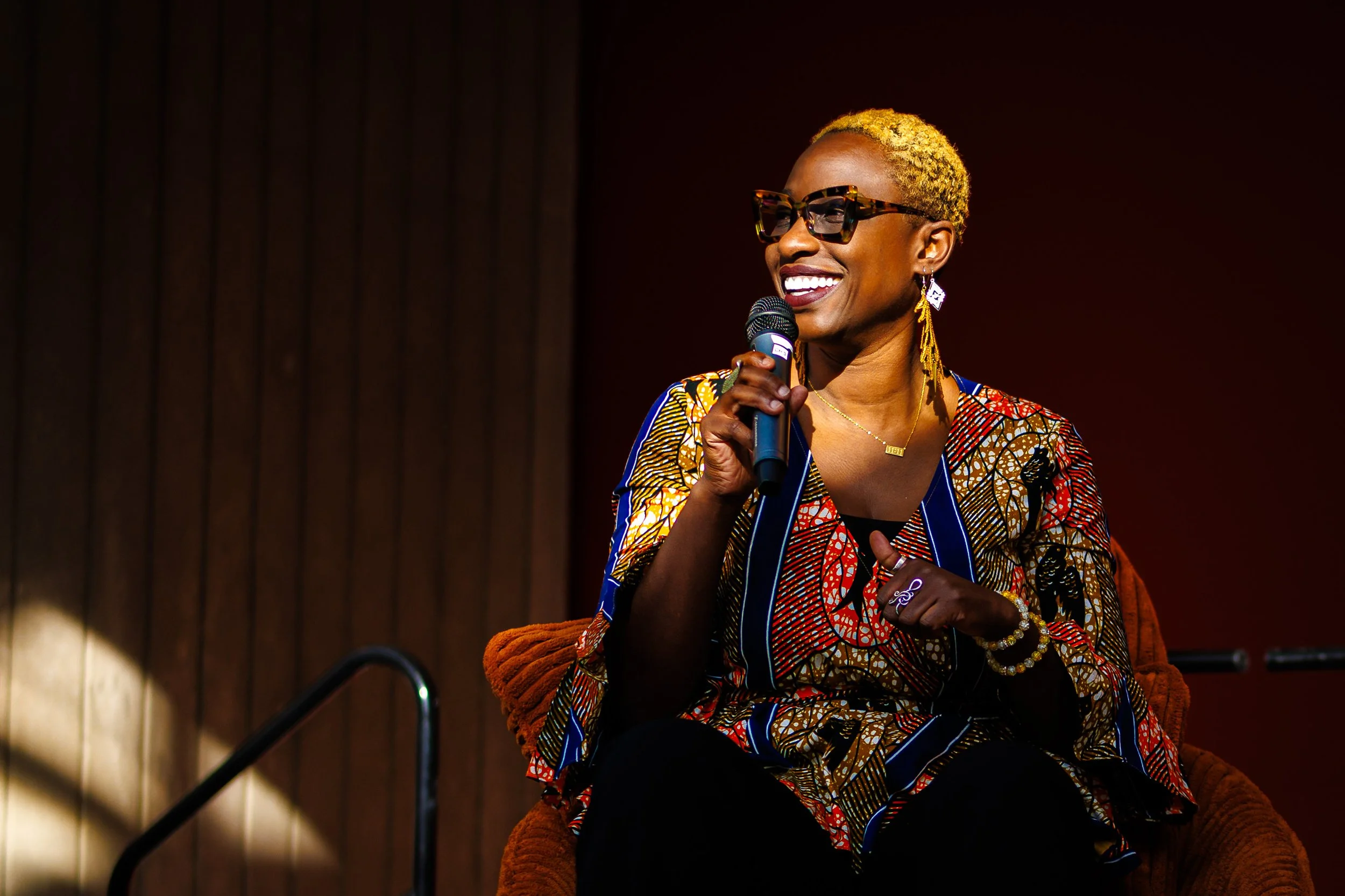 Black woman with blond hair holds a microphone sitting in an orange chair